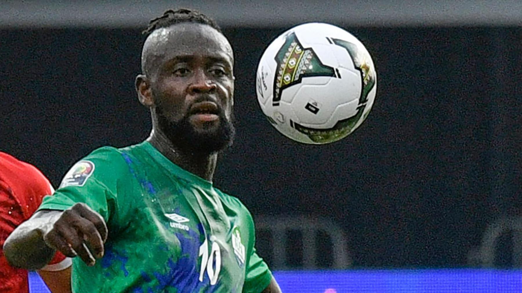 Kei Kamara, pictured from the chest up wearing a green Sierra Leone shirt with the number 10 on the chest, looks to control a football which is bouncing in front of him at head height