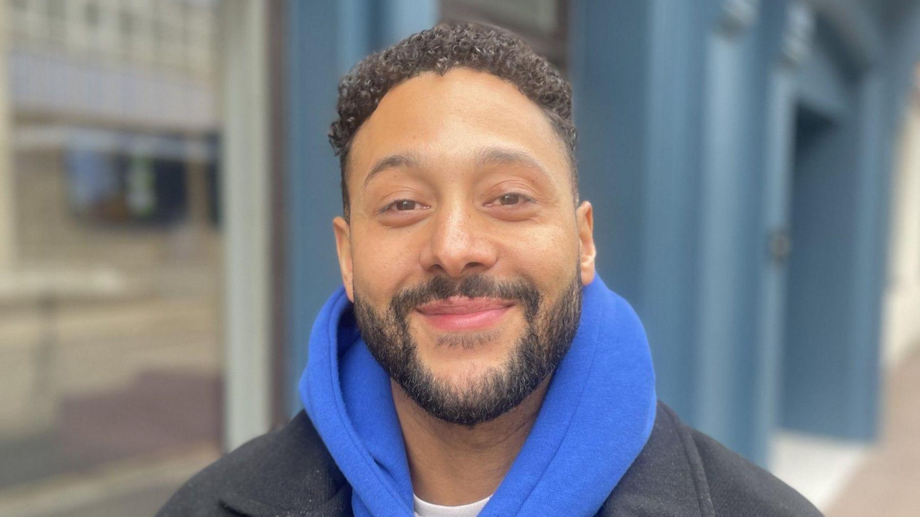 A man with a black beard and hair. He is wearing a blue hoodie and smiling. 