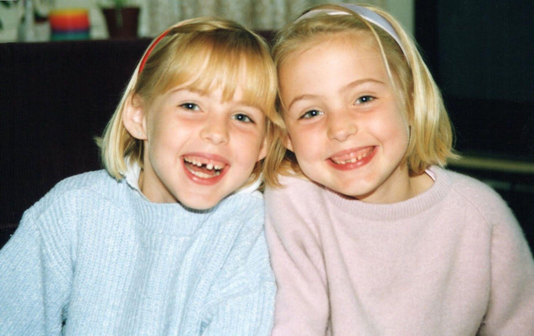 A picture of Kris and Maren as young children. They have shoulder length blonde hair and are wearing pink and blue jumpers. They are smiling at the camera.