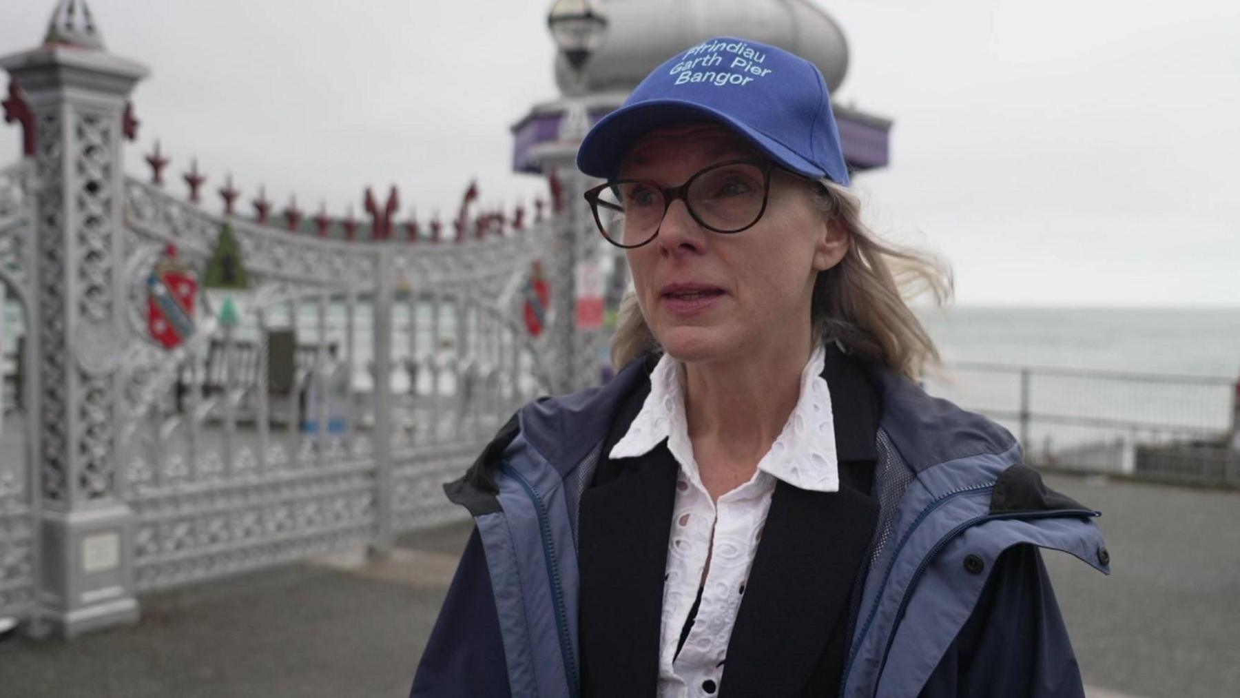 Vanessa Hawkins who is wearing a blue baseball cap and large glasses and has blonde shoulder length hair. She is wearing a blue raincoat over a black blazer over a white shirt