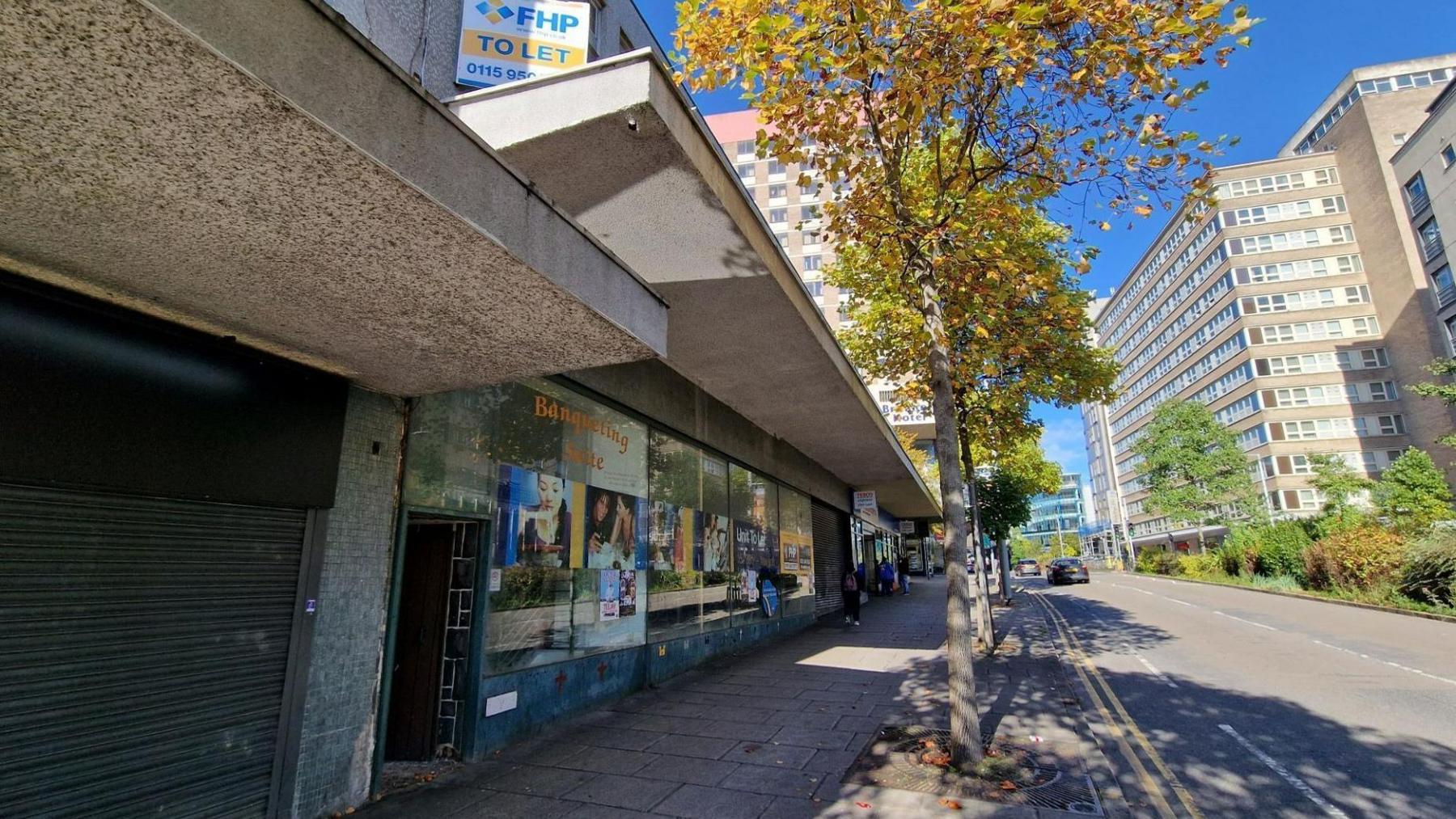 A view of a wide urban street with a concrete shop front, and the large windows have old posters for nearby attractions