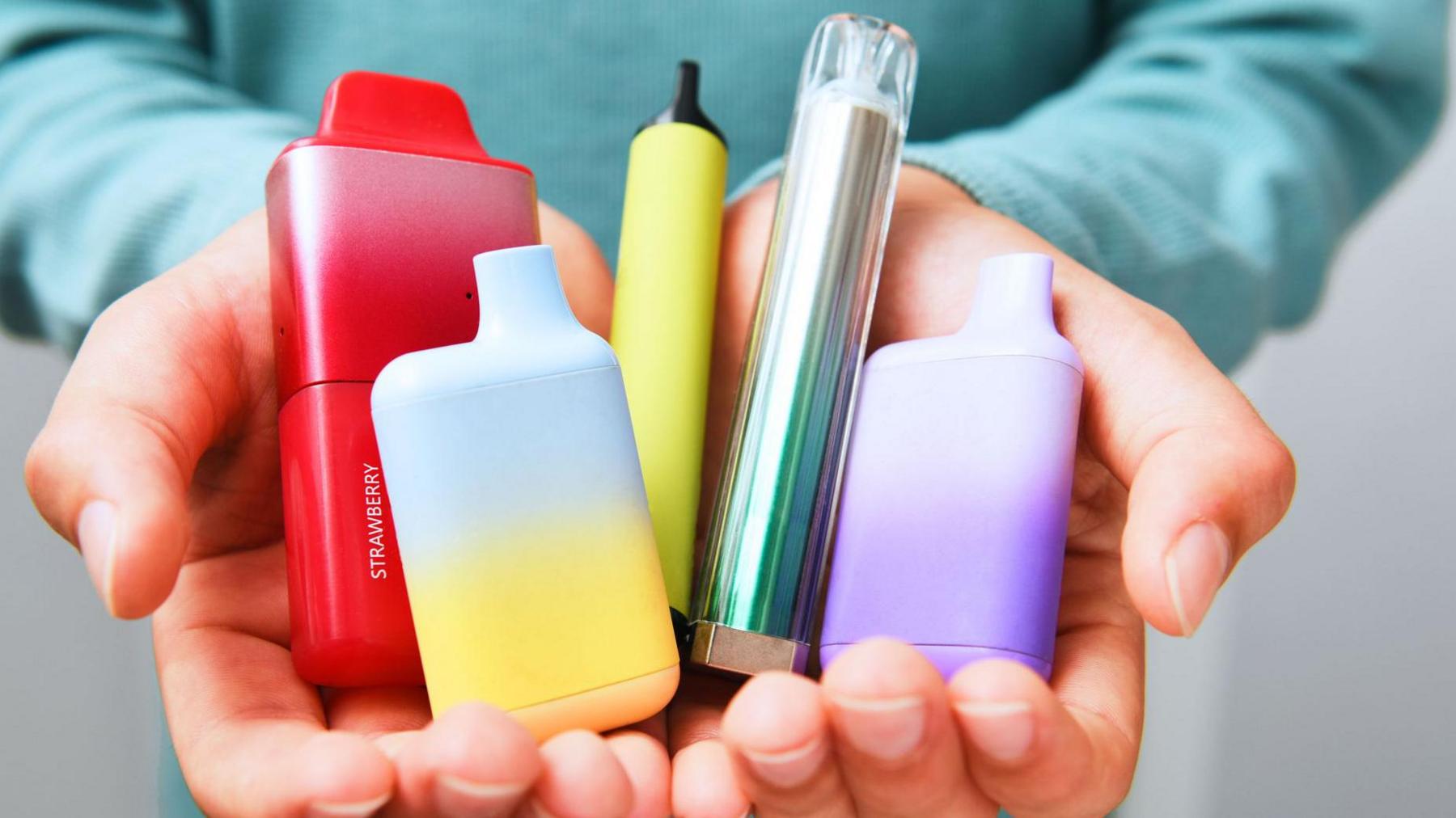 A person's hands with them showing vapes, including a red, large vape which says "strawberry" on the side and a yellow and blue vape.