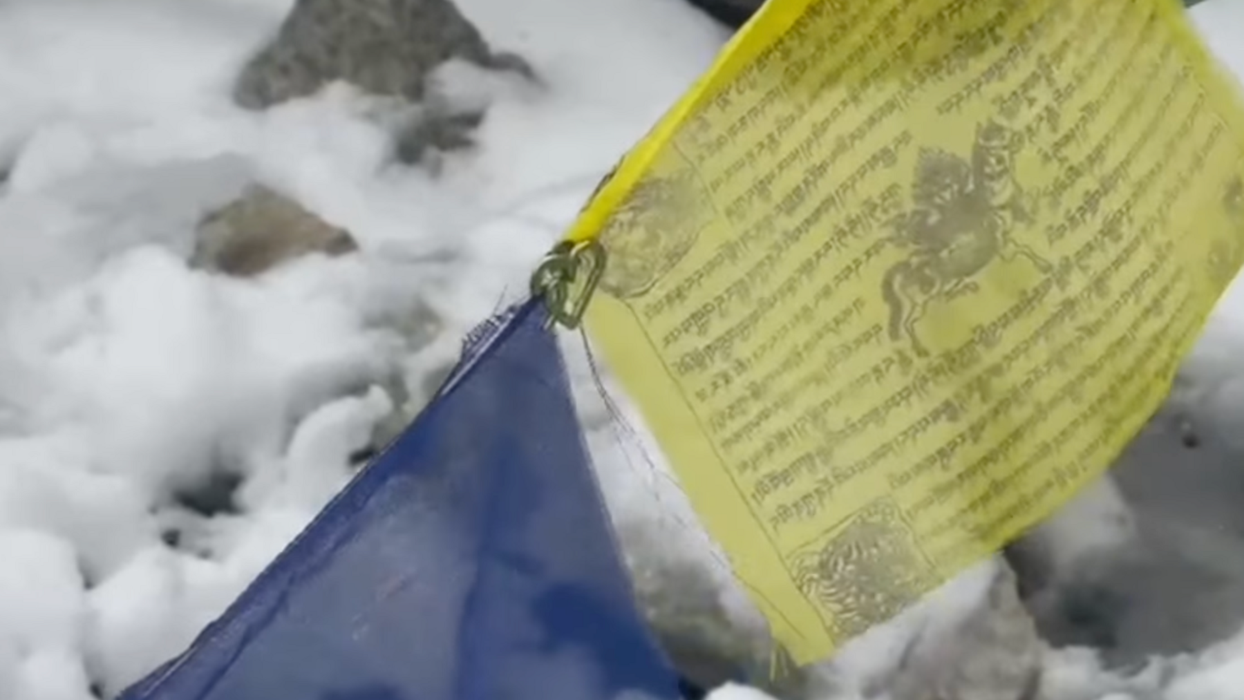 A small green metal heart is clipped onto some string that holds up a yellow and blue flag. The yellow square flag has writing on it in a different language and the symbol of a horse. There is snow in the background.