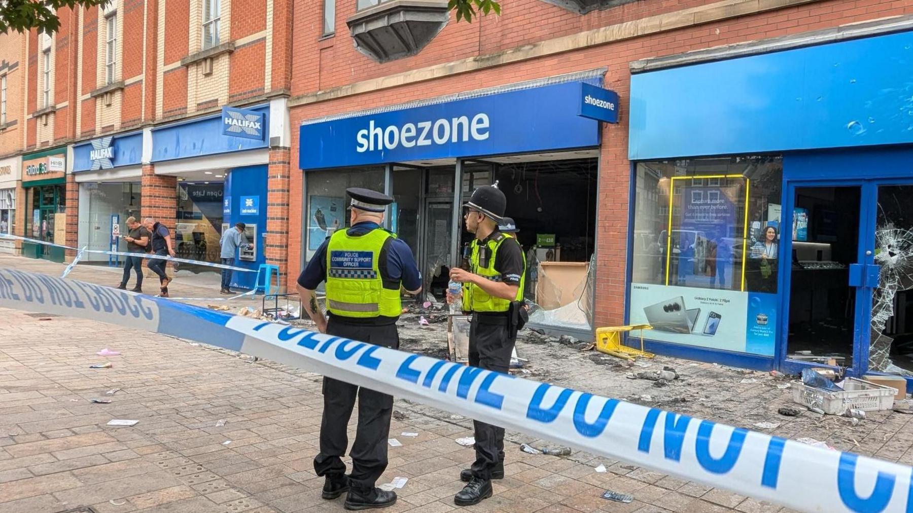 Two police officers stand outside Shoezone and the O2 shop in Hull city centre behind police tape. The aftermath of the looting can be seen with broken doors and windows and the remnants of a fire in the background.