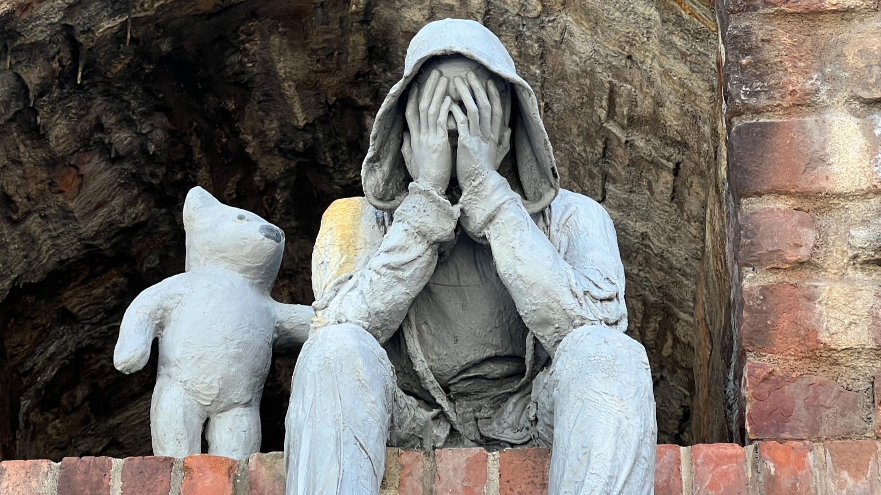 A close up view of a grey sculpture depicting a seated, hooded male figure with hands covering its face, accompanied by a bear-like companion offering comfort with a paw on the figure’s shoulder. 