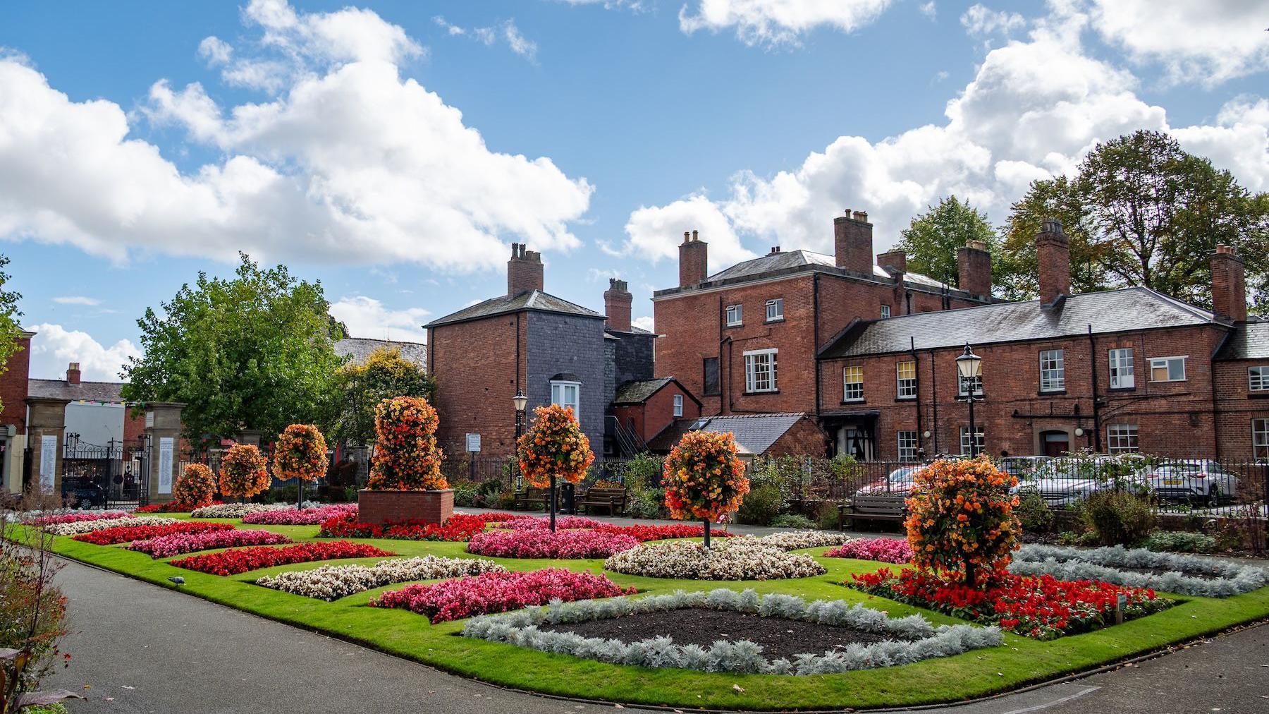 A section of green grass in a park surrounded by a concrete path. On the grass are flowerbeds featuring rectangles of pink, red and white flowers. There are orange bushes across the length of it, increasing in height.