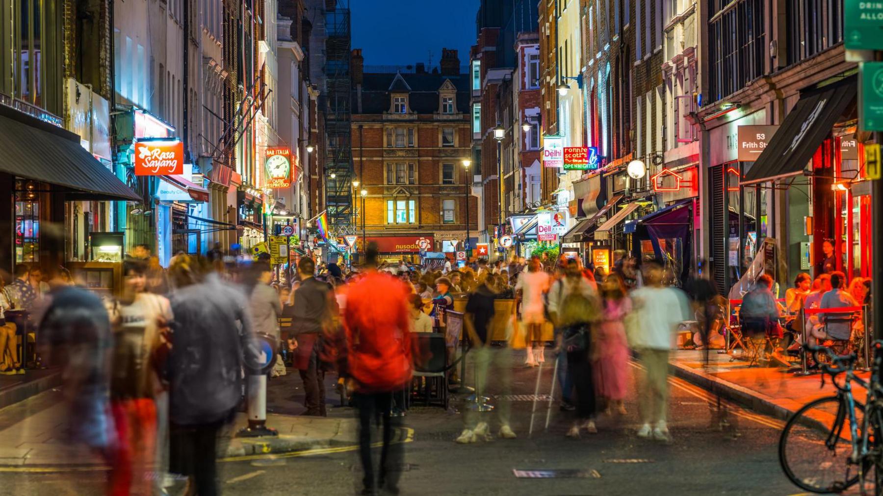 A blurred image of a busy street in Soho at night. There are neon signs outside businesses on either side of the street, and the street is full of people.