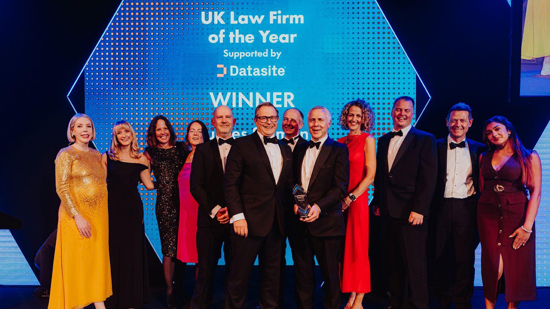 A group of people - a mixture of men and women - stand in a group facing the camera. They are all from law firm Burges Salmon. The men are all wearing black tie and the woman are in smart dresses. They are in a dark space illuminated with blue and white light
