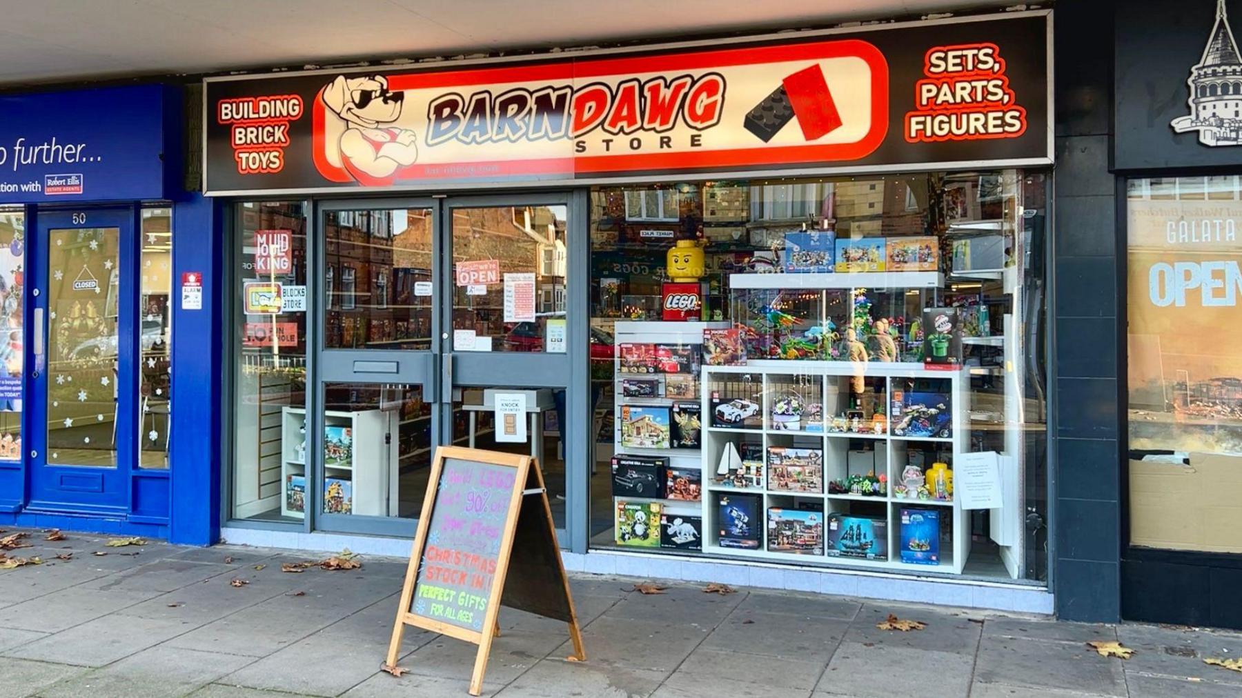 A glass-fronted shop with a large red, white and black sign which reads "BarnDawg Store." The window is filled with Lego models. There is a chalk A-board outside the shop. The shop is attached to a blue shop to the left and a black shop to the right.