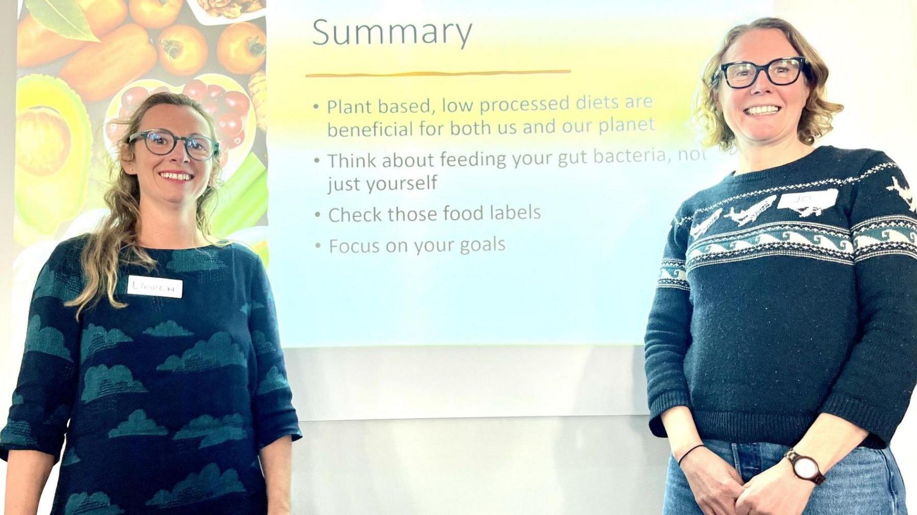 Two smiling women dressed in a top with clouds on it and a blue top with waves on it stand in front of a slide with tips on to to eat healthily.