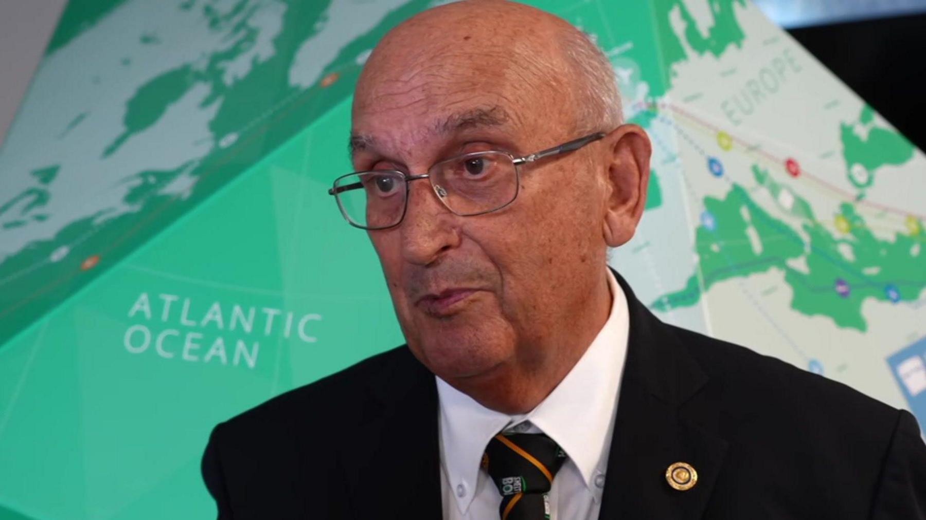 A man is dressed in a dark suit and a gold and black tie. He has a Rotary pin in his jacket collar.
He is standing in front of a green globe.