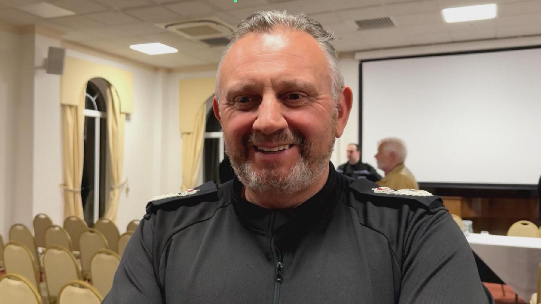 middle aged man with grey hair and a black shirt he is in a room with rows of chairs behind him.