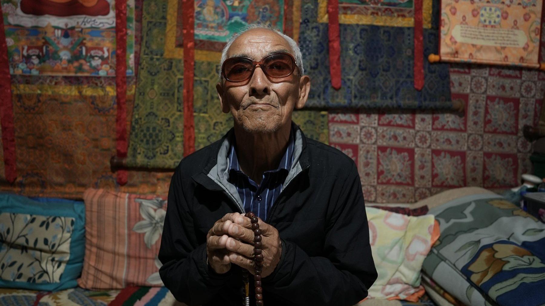 Dawa Sangbo with sunglasses sits, wearing a black jacket over a blue striped shirt and holding prayer beads. The wall behind him is covered in colourful rugs.