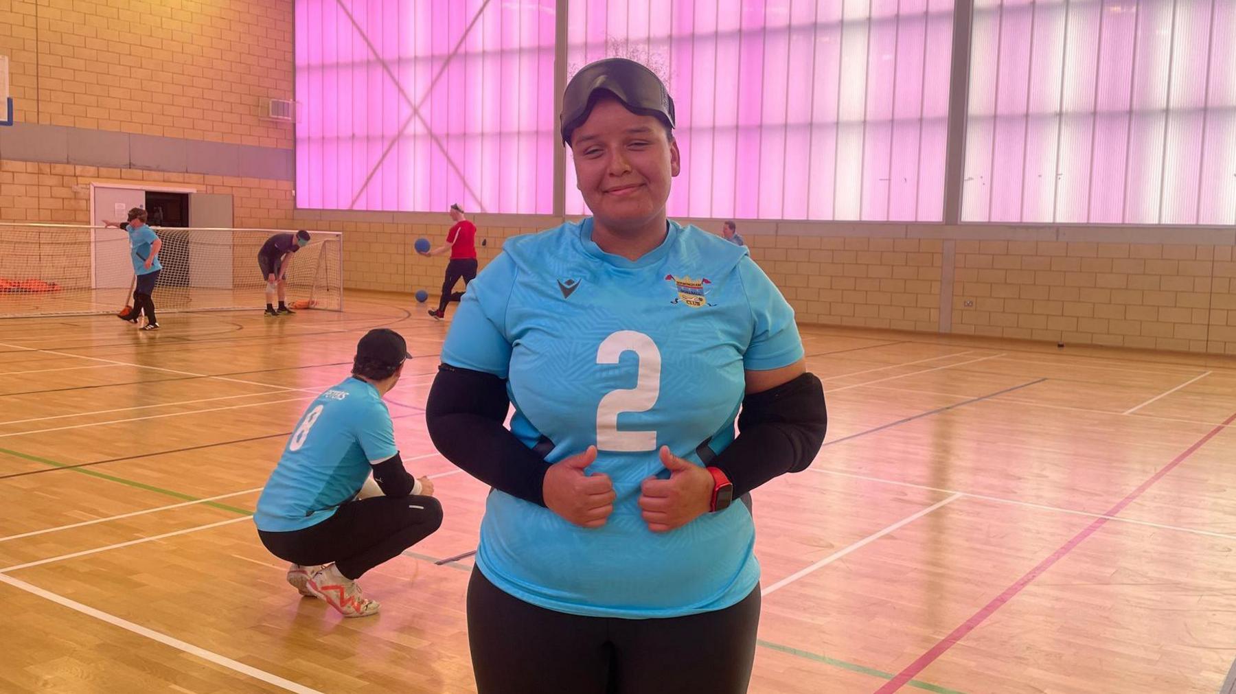 Goalball player Alex Ulysses is wearing a blue sports top with the number two in white on the front of it. She is stood in a sports hall with other players in the background. There are a set of goals and a wall which is illuminated with purple light.