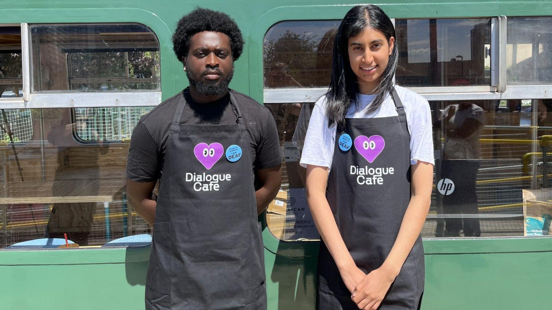 Two young baristas, wearing black aprons with the Dialogue cafe logo on the front - which includes text in white and a large purple heart - stand in front of the café's windows.