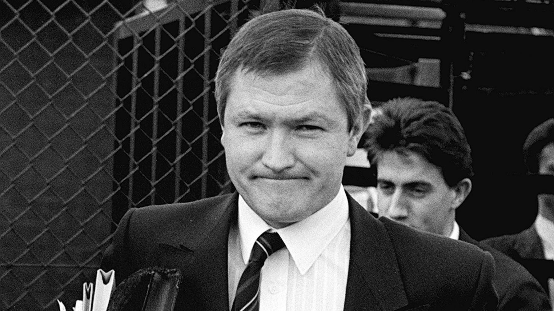 A black and white image of solicitor Pat Finucane standing outside of a court building. He is carrying a bundle of papers and files and wearing a black suit jacket, a white collared shirt and a tie.