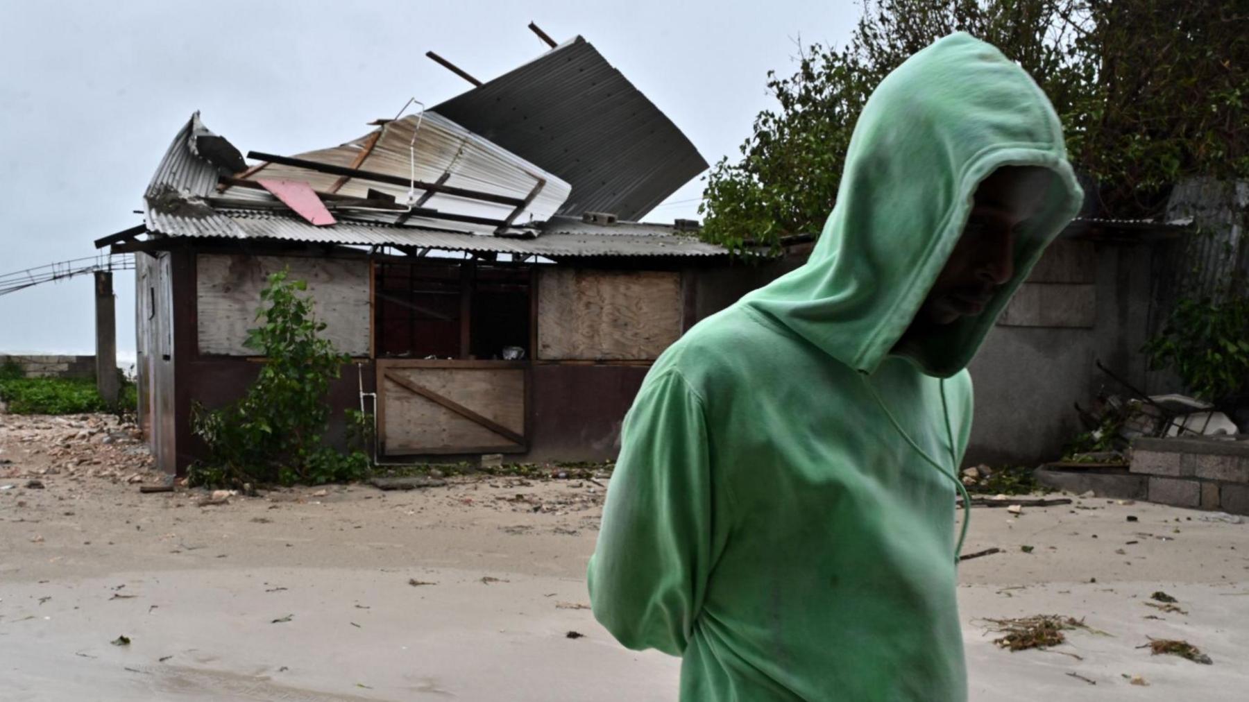 A man in a green hoodie walks past a damaged property in Jamaica