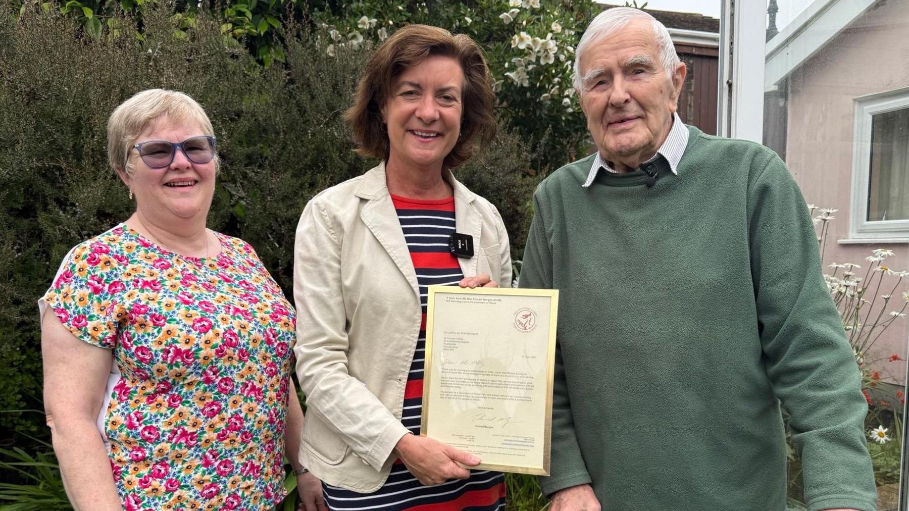 The First Minister of Wales presents Duncan Hilling with a letter of thanks in a gold frame with his daughter, Miranda, at the presentation in his garden. Miranda is on the left and wearing a pink and yellow flowery top. She has light blue fluorescent shades on and is smiling. She has short blonde hair. Eluned Morgan is in the middle, and has dark-brown hair in a bob-style. She has a cream jacket over a blue, red and white striped dress. Duncan is at the end and is wearing a green jumper with a white collared shirt, the shirt collars are visible. He has short grey hair. 