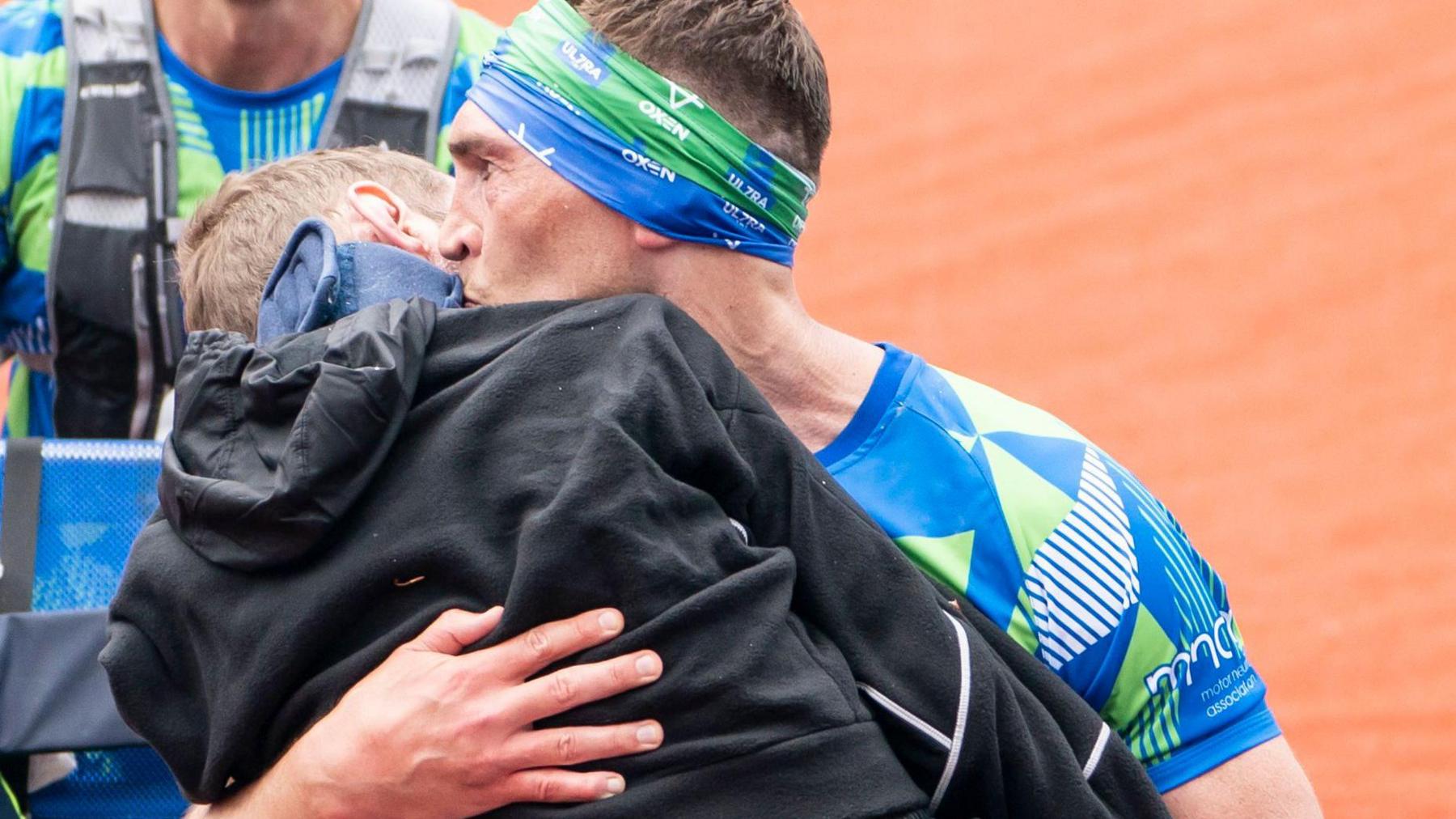 A man in a blue and green patterned top and head band carries a man wearing a black top in his arms and kisses the side of his face. Behind them is another runner in a green and blue top.