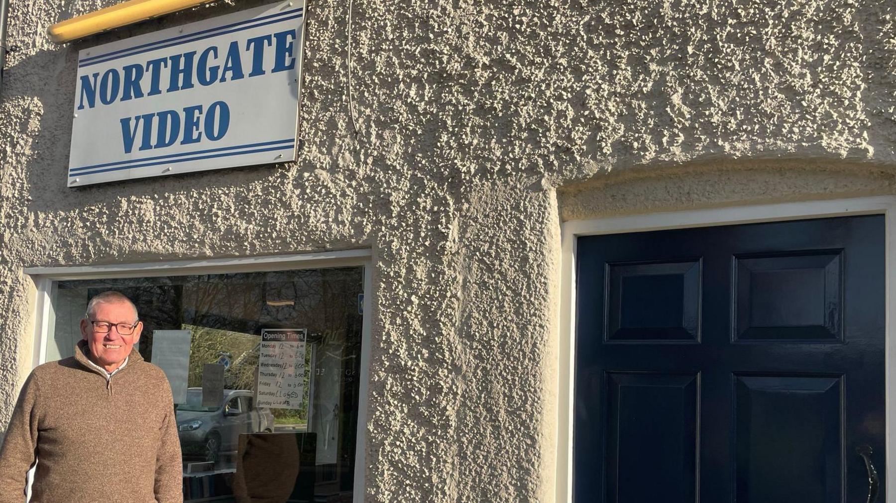 Steve Webber stands outside the white building. It has a black door. He is smiling in the direction of the camera .There is a large window behind him, through which you can see DVDs. Above the window is a white sign with blue writing that reads 'Northgate Video'.