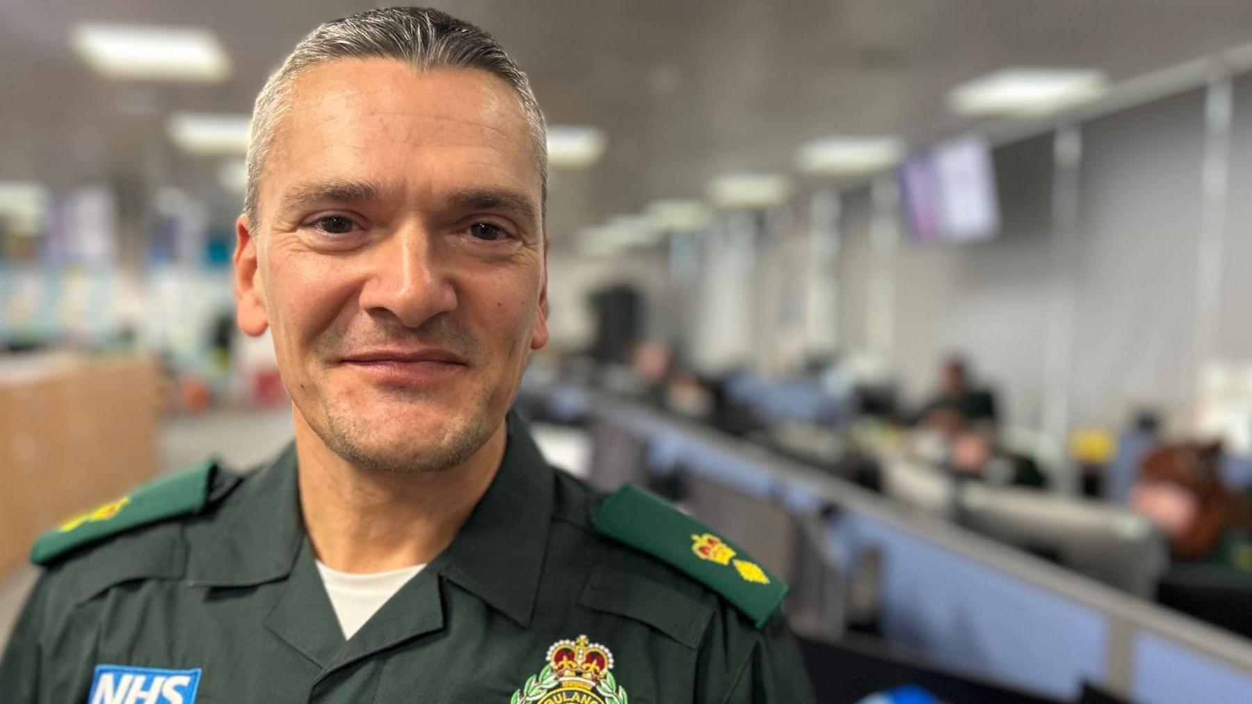 Stuart is pictured wearing his dark green North East Ambulance Service uniform. He has black and grey hair and is clean shaven. 