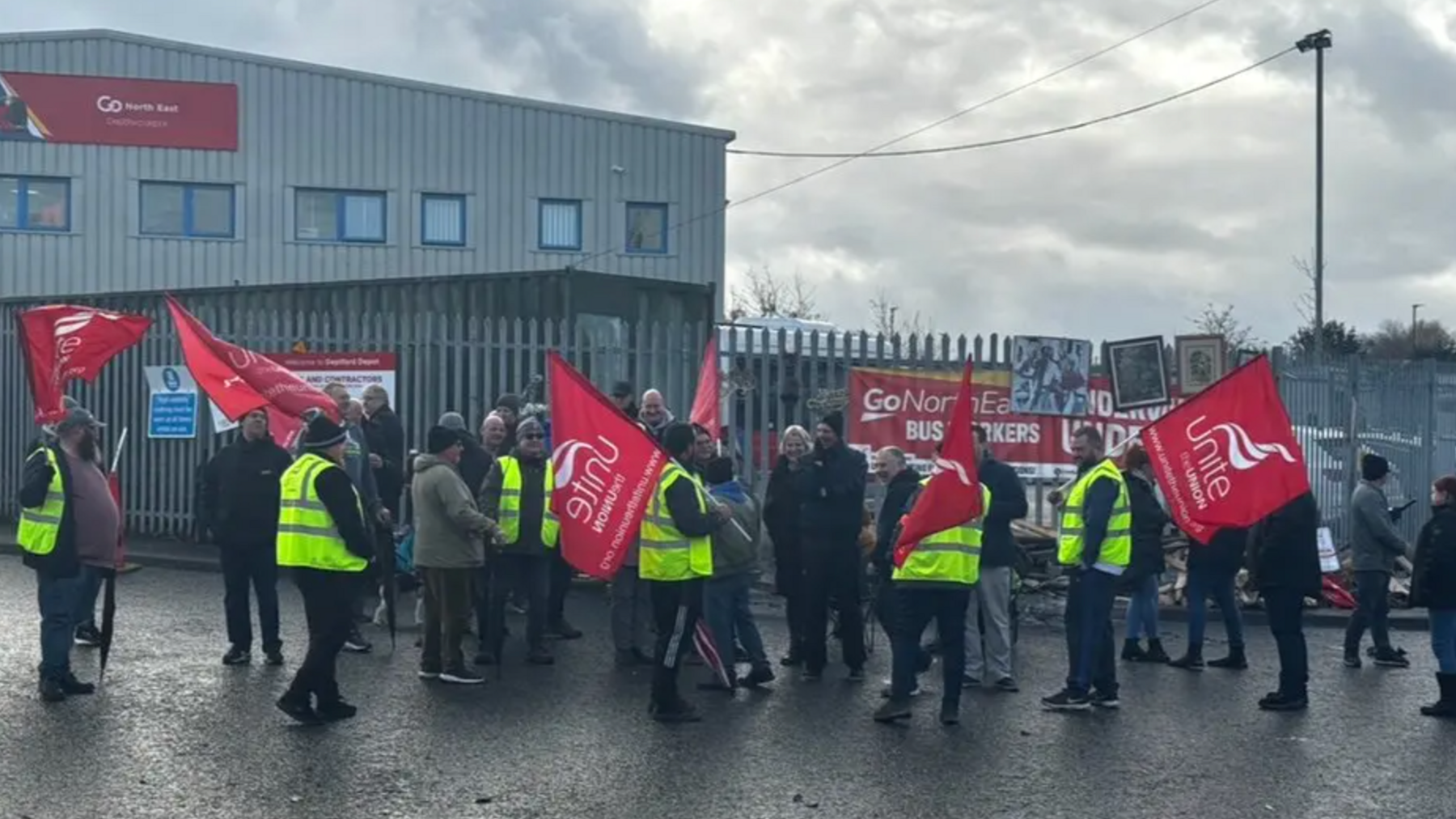 A picket line outside Go North East's depot. People in hi-vis vests stand in the cold holding red Unite union flags. 