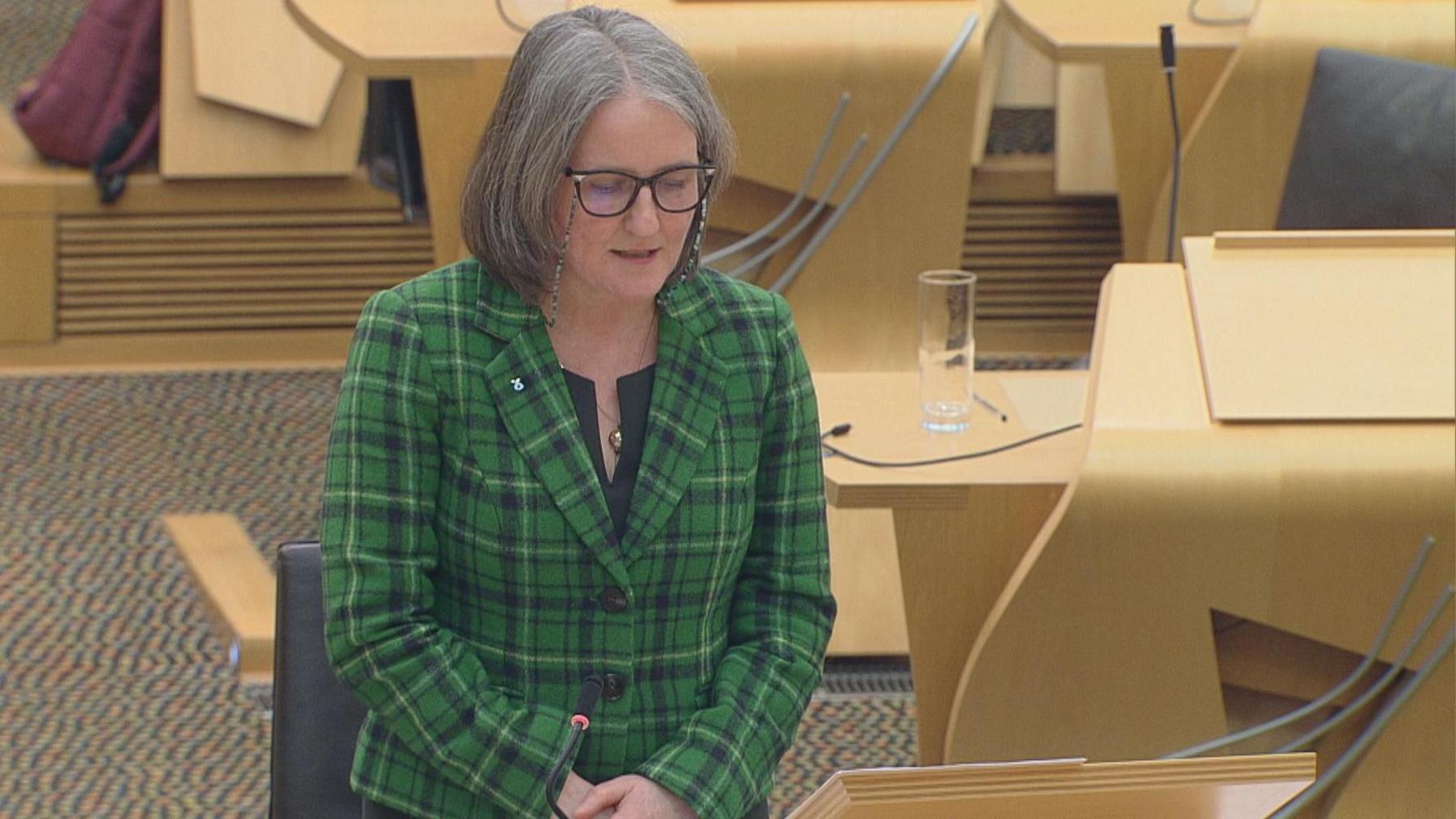 Maree Todd wears a green tartan jacket, black top, silver necklace and black-framed glasses with a retainer chain. She is standing in the chamber in Holyrood, with wooden desks and black seats behind her.