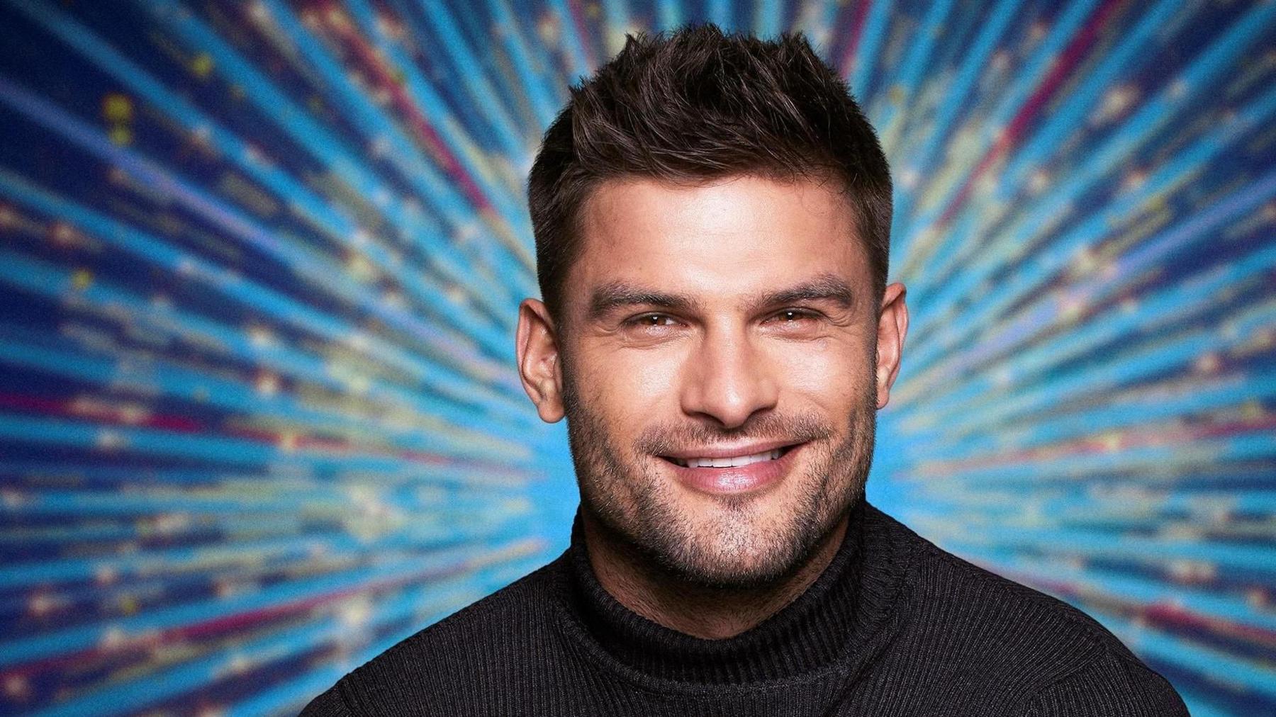 Aljaz Skorjanec faces the camera wearing a black turtle neck jumper, smiling. He has spiky dark hair. Behind him is a blue starburst image