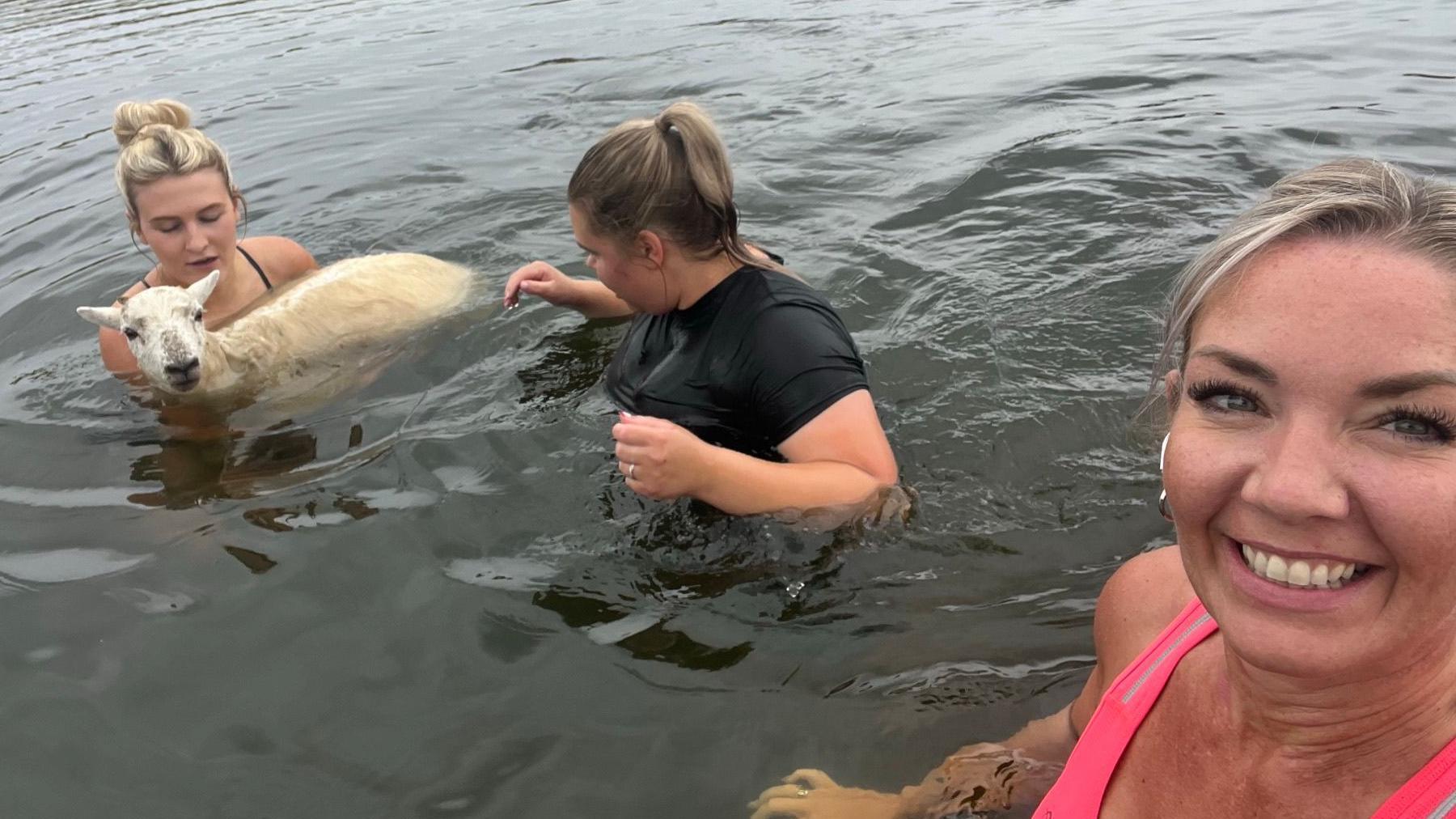 Aimee Turpie, who is wearing a pink running top smiles broadly as she takes a selfie to show Britney Lawrence and Abi Cox rescuing a sheep in Siblyback Lake in Cornwall. Britney is carrying the sheep in the water. Abi is stood next to her and is wearing a black top. All three are either waist or chest deep in the water.