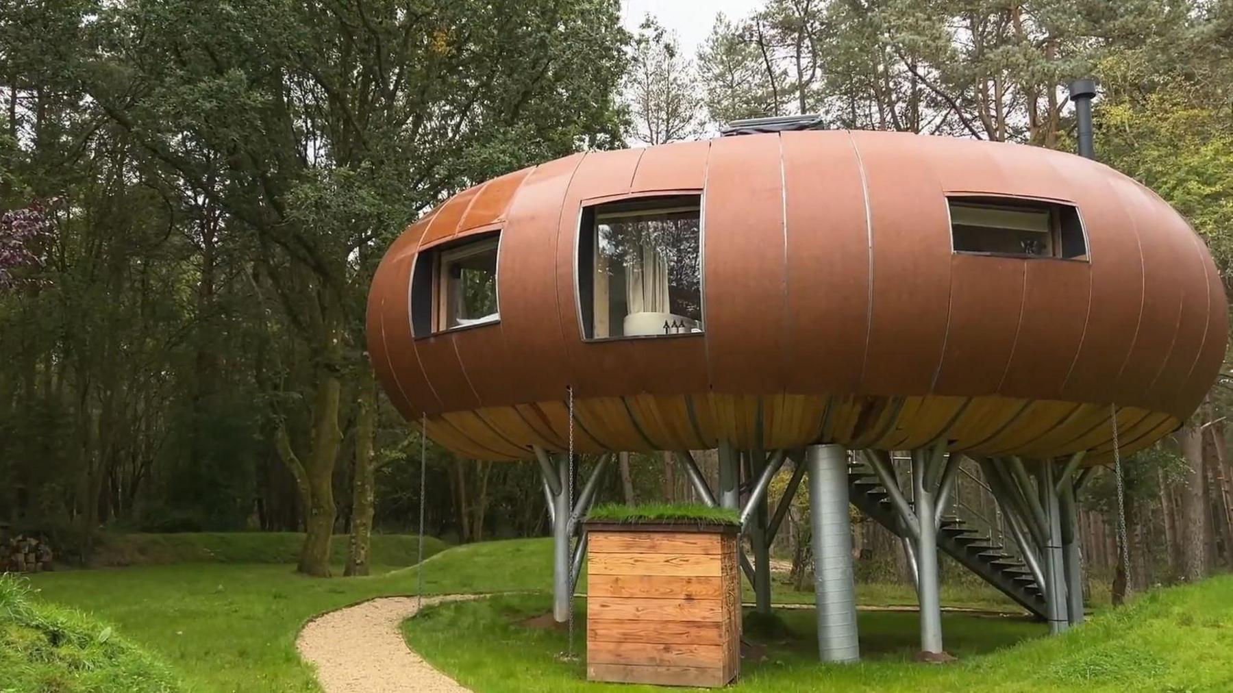 A large brown ufo-like treehouse. It is elevated off the ground by metal stilts and is surrounded by trees.