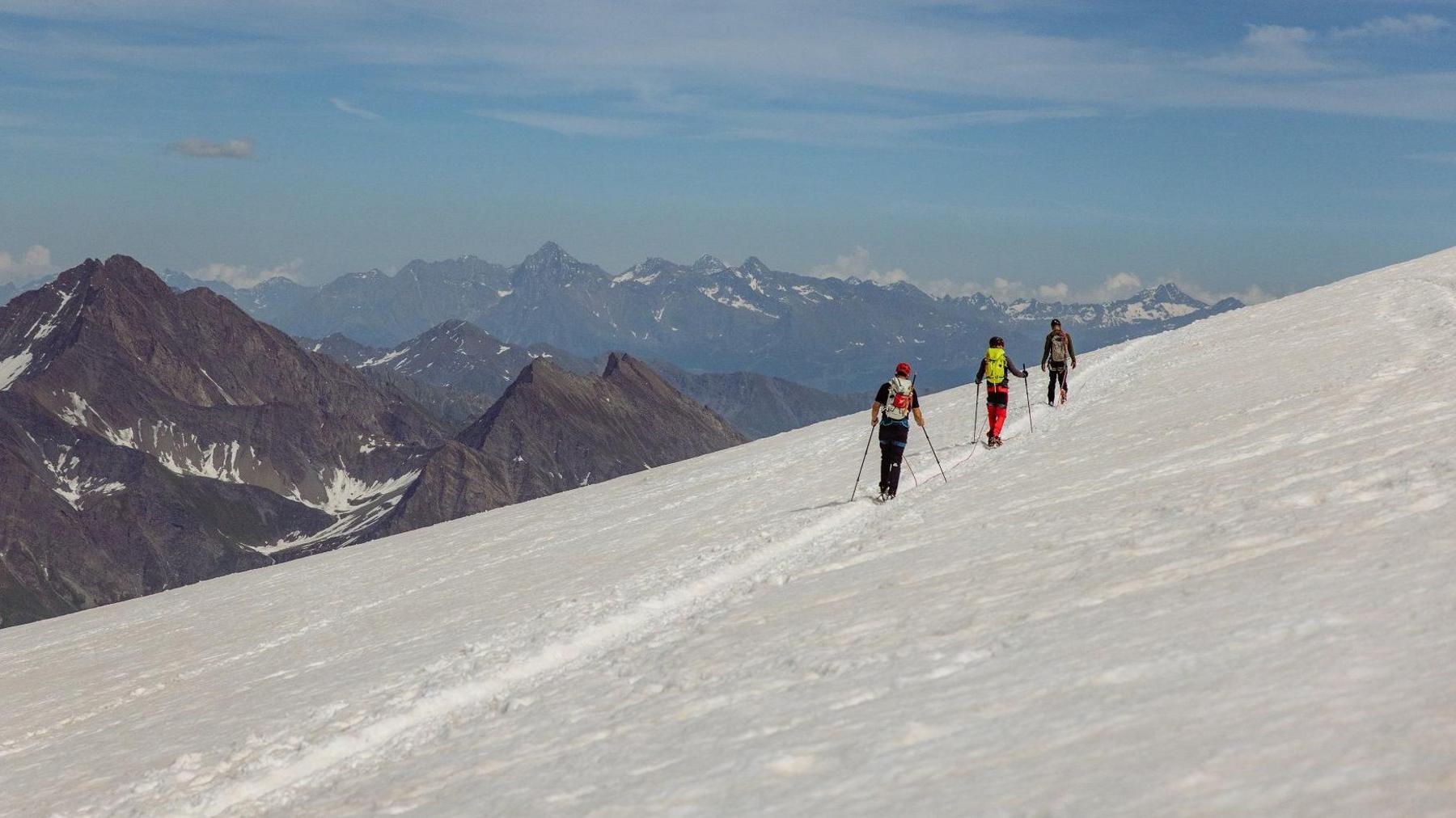 Three hikers with backpacks and poles trekking along a snow-covered mountain trail, with rugged peaks and a partly cloudy sky in the background.