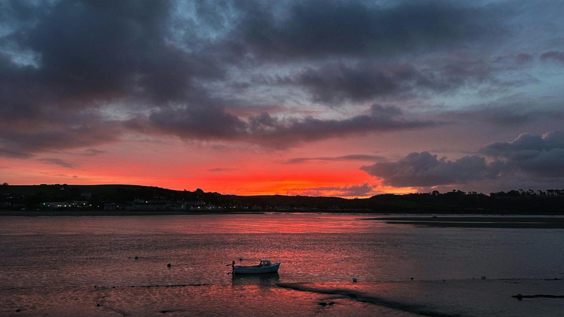 A stunning sunrise can be seen above Appledore. Liz took the photo looking out across the water looking towards the town. There are grey clouds above the scene and shades of orange and pink breaking through the gloom. A small boat can be seen moored in the water in the foreground.