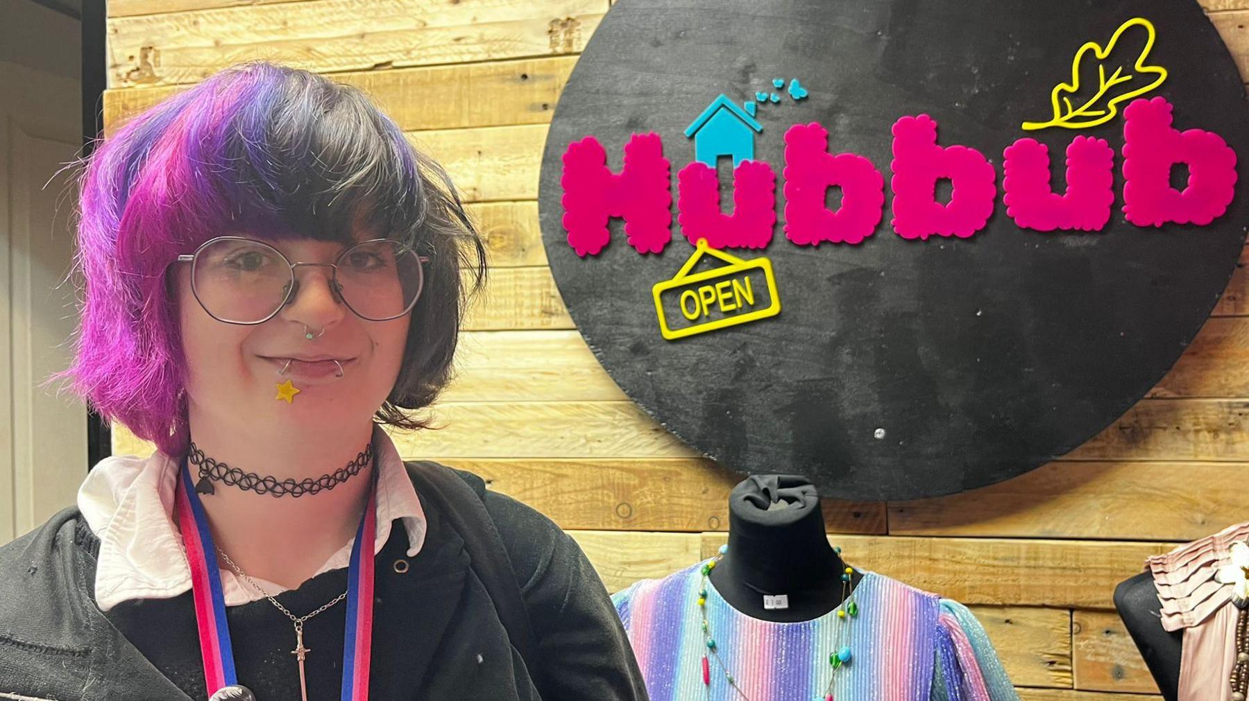Person gyda gwallt piws a sbectol crwn yn gwenu i'r camera, o flaen logo pinc a du yn dweud 'Hubbub'.
