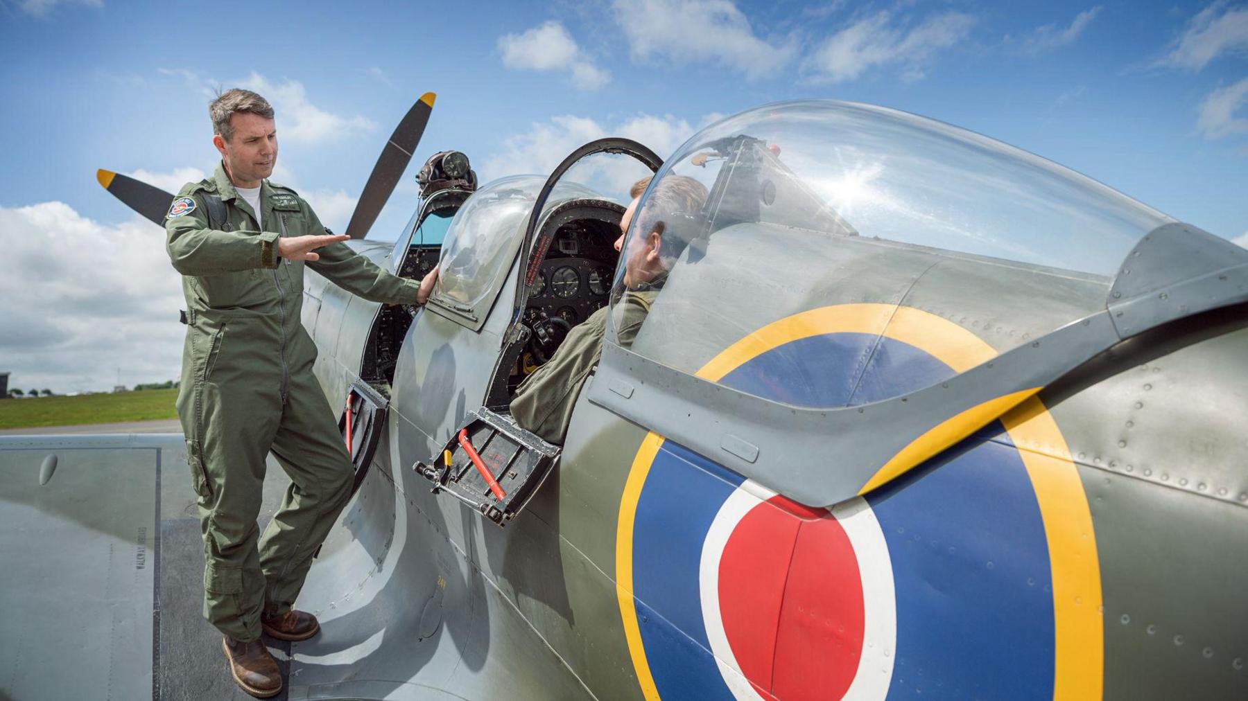 A pilot standing on the wing of an aircraft speaking to someone in the passenger seat, which is behind the pilot's seat in the cockpit.