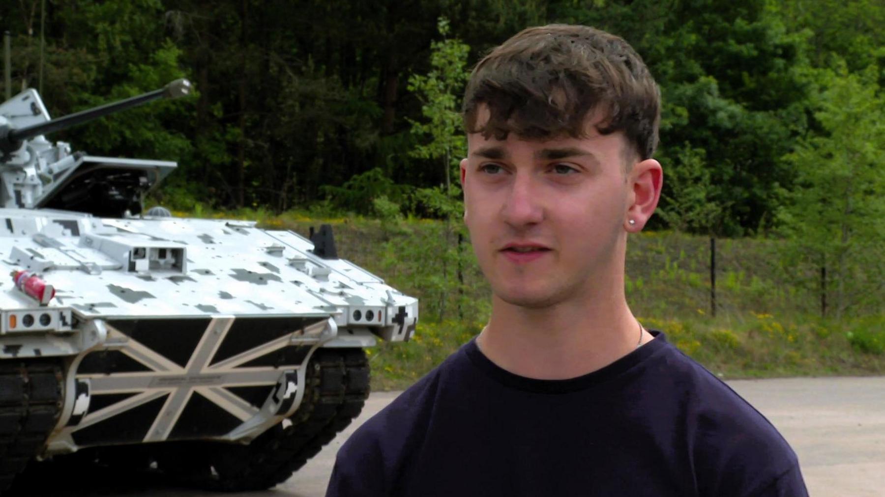A dark-haired man in a navy crew-neck tshirt stands to the right of an armoured vehicle 