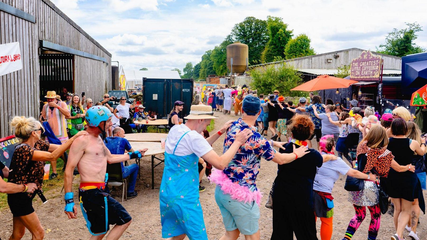 A group of people doing the conga at a festival. Many of the people are dressed in colourful festival clothing.