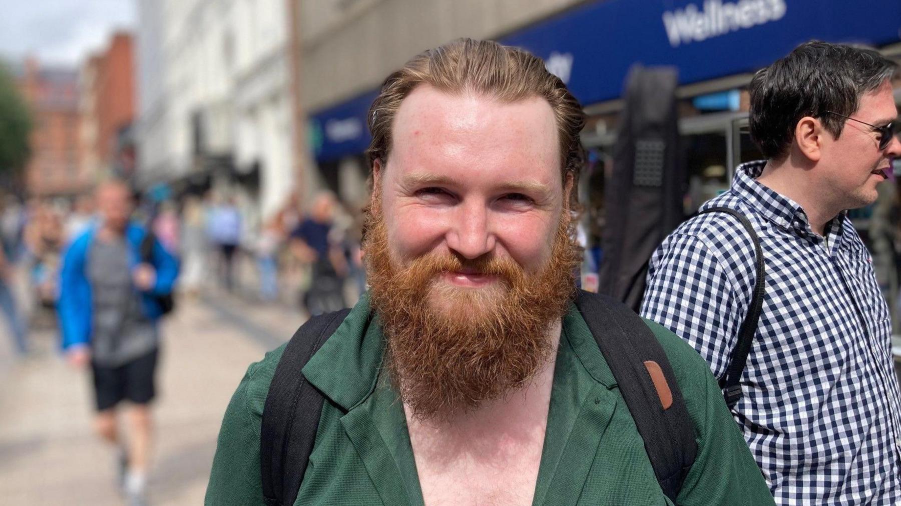 A man smiles at the camera. He has an auburn beard and is wearing a green shirt. He is standing on a busy street.