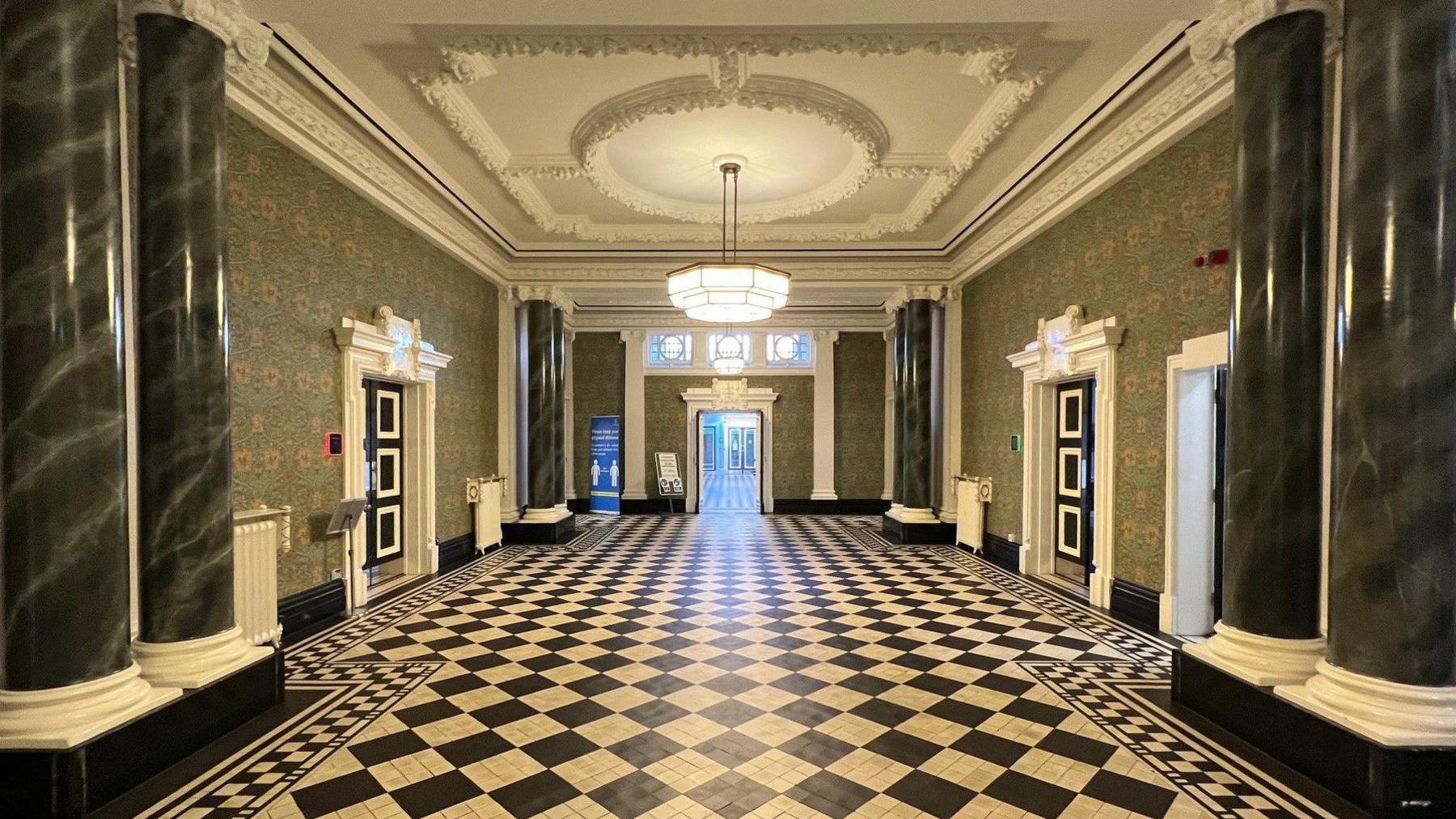 A large hallway with black and white tiled floor and darkly-coloured columns from floor to ceiling, with ornate decorative features above doorways and on the ceiling. There are doors either side of the hallway, and a doorway leading into another corridor directly in the centre of the image.