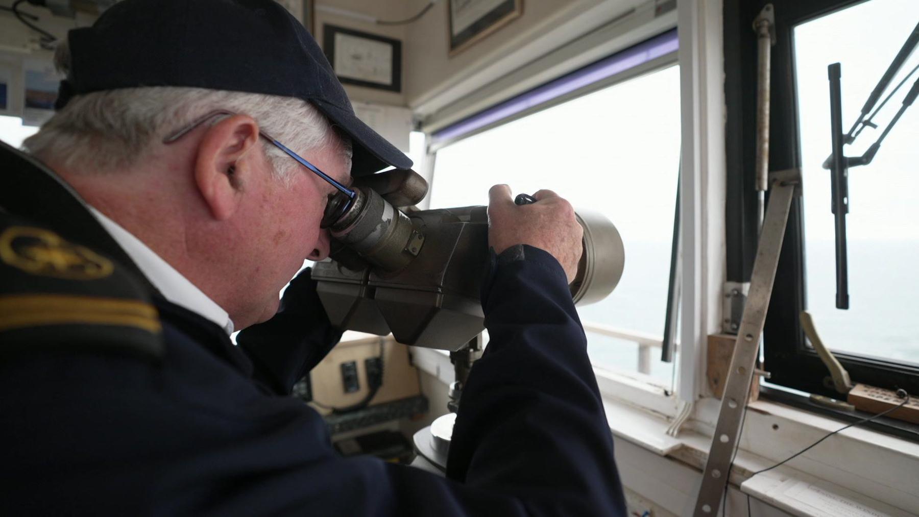 Jerry wears a dark cap and top with gold epaulettes. He is looking through a telescope through a window out to sea.