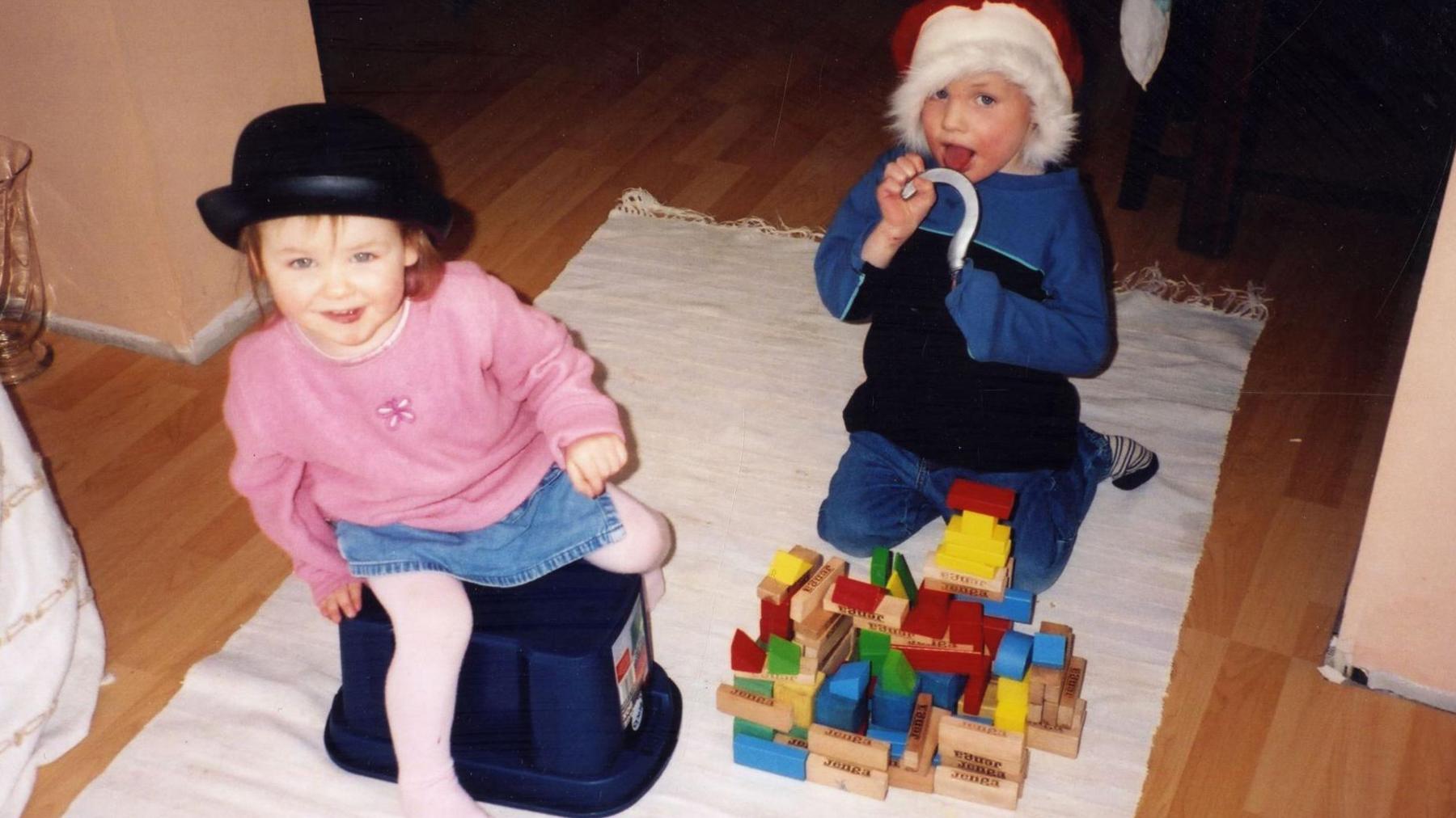 Hannah is wearing a bowler hat and is sitting on an upturned toy box. Andrew is wearing a Santa hat and is sticking his tongue out. There is an assortment of wooden building blocks on the floor.