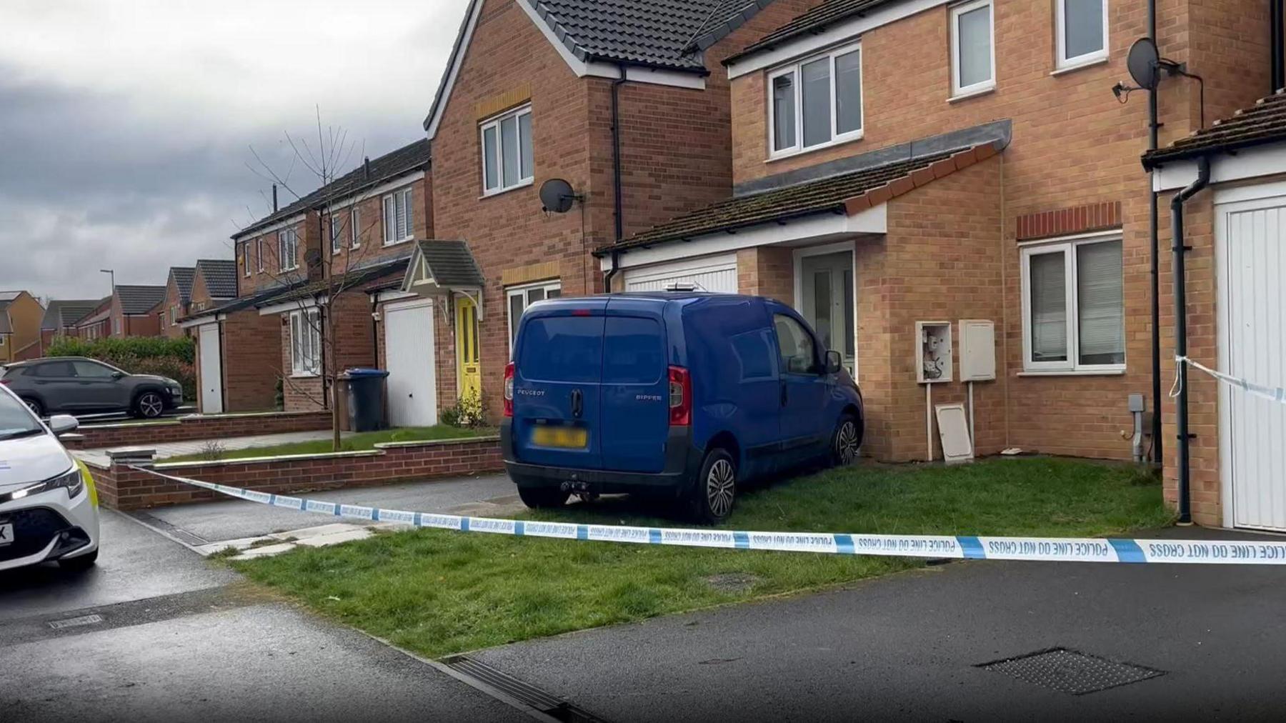 A small blue van has its bumper against the porch of a house behind a police tape.