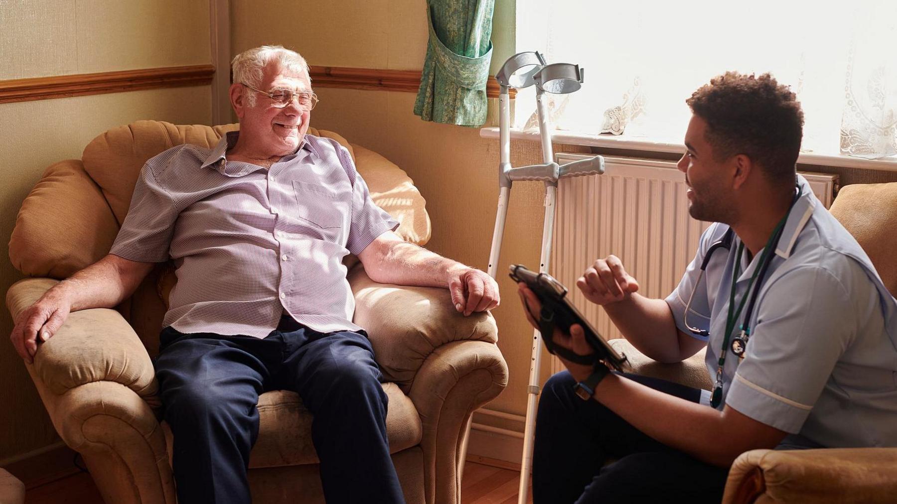 Older man lounging in armchair with crutches to one side, young male nurse seated in another chair filling in form on a tablet