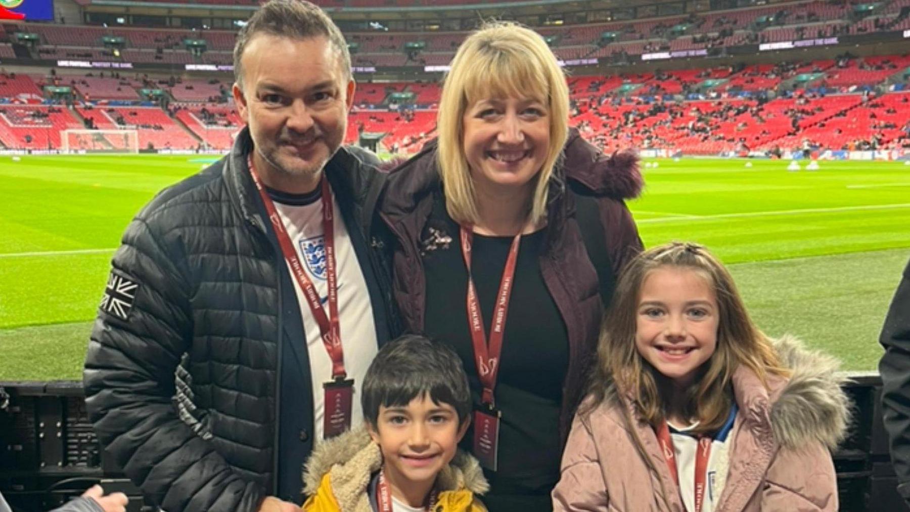 Lee Rawlinson and his wife and children.
The man is on the left wearing a black puffer jacket and is standing next to his wife who is blonde and wearing a black coat.
In front of them are their children.
A boy about 6 wearing a yellow coat and a girl about 10 wearing a light pink coat.
They are at Wembley. 