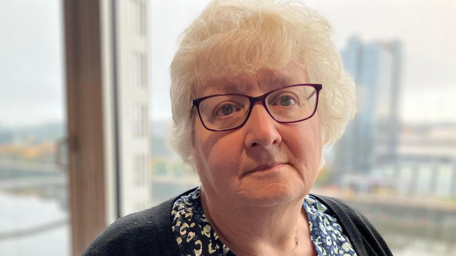 Laura Schosloff is staring at the camera. She has grey hair and glasses and is wearing a patterned black and white blouse and a black cardigan