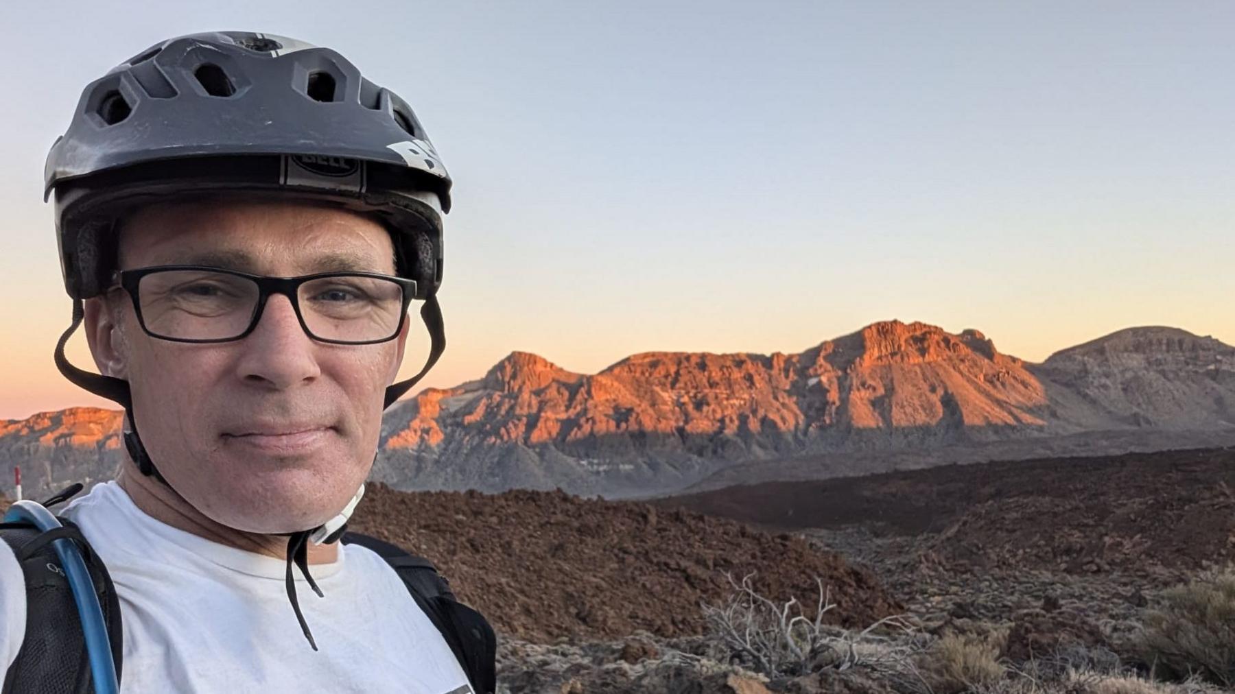 A man wearing glasses, a black cycling helmet, white t-shirt and a black rucksack is standing in from of a dormant volcano at sunset. The tips of the peak are touched by the orange glow of the sun.
