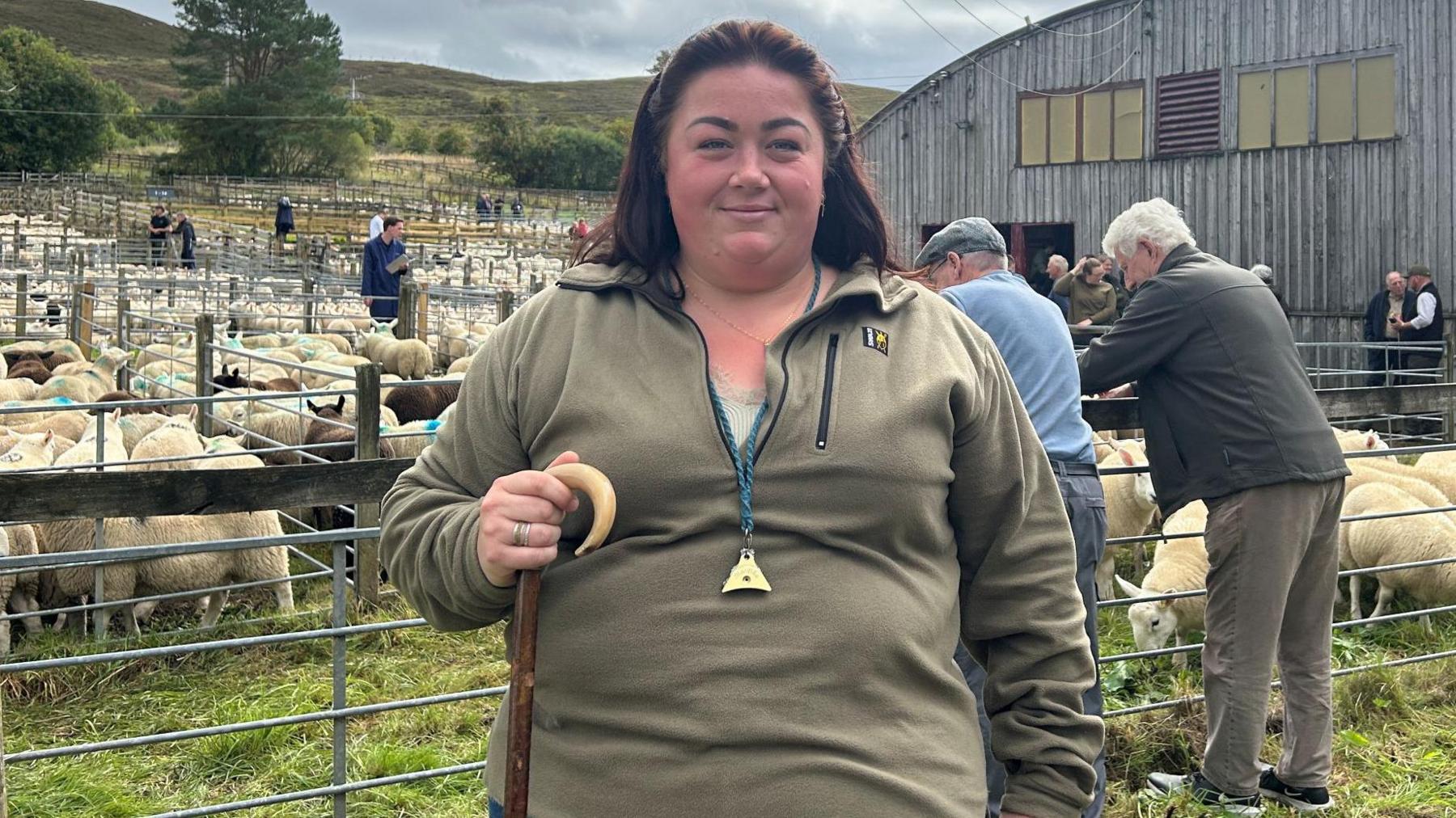 Shepherdess Cara Lowe is standing outdoors at the Lairg lamb sales. She has long brown hair and is wearing a light green fleece. She is holding a shepherd's crook and has a whistle to instructing sheepdogs hanging from a lanyard around her neck. 
