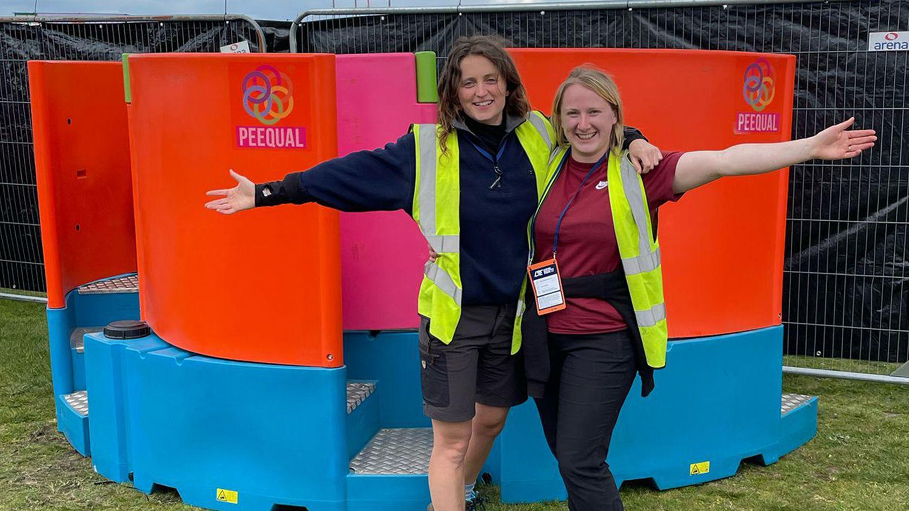Two women stand with one arm around each other and the other outstretched to their side while smiling at the camera. They are both wearing yellow hi-vis vests and are standing in front of their design for a women's urinal. It is orange and blue in colour and has the word Peequal written on it