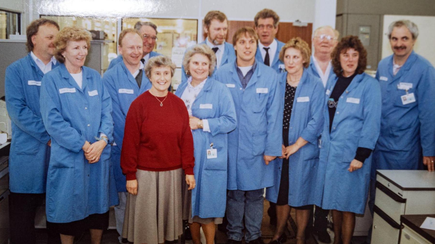 A photo from the mid-1990s shows a middle-aged woman wearing a red jumper and a brown skirt smiling. She is surrounded by workers in blue coats standing in an open office area, with a man on the right leaning on a filing cabinet.