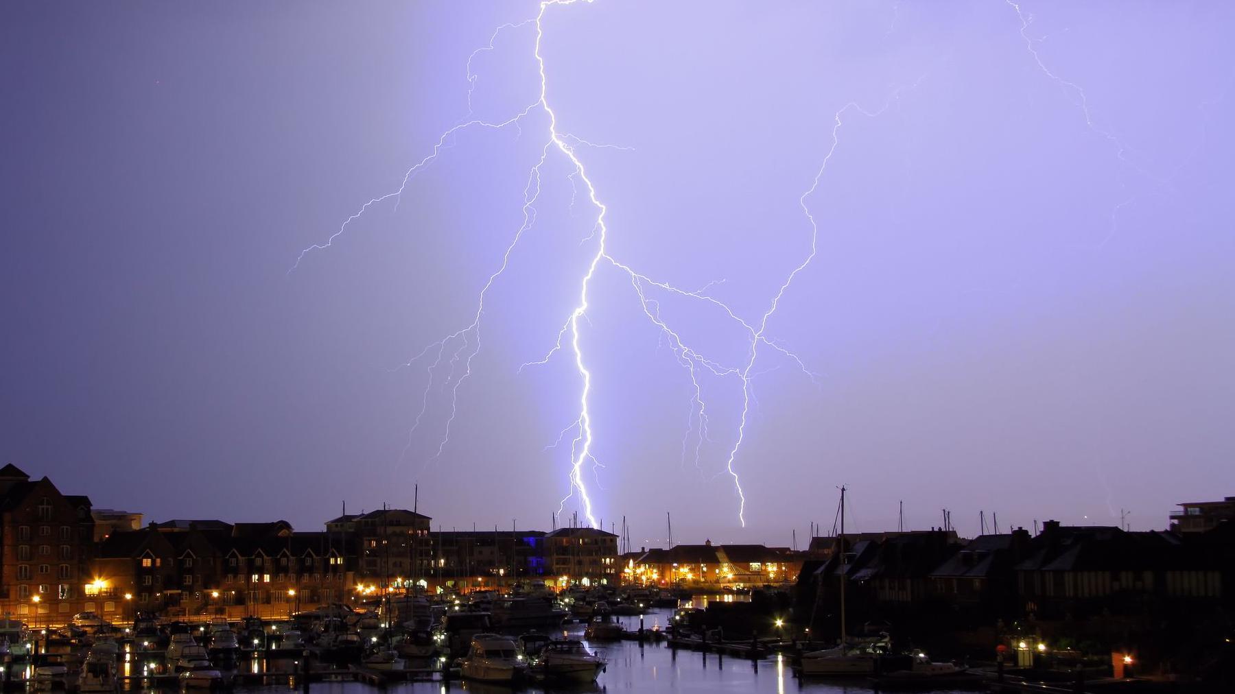 A fork of lighting flashes over Sovereign Harbour, Eastbourne, at night. Boats and the seafront buildings in the foreground.