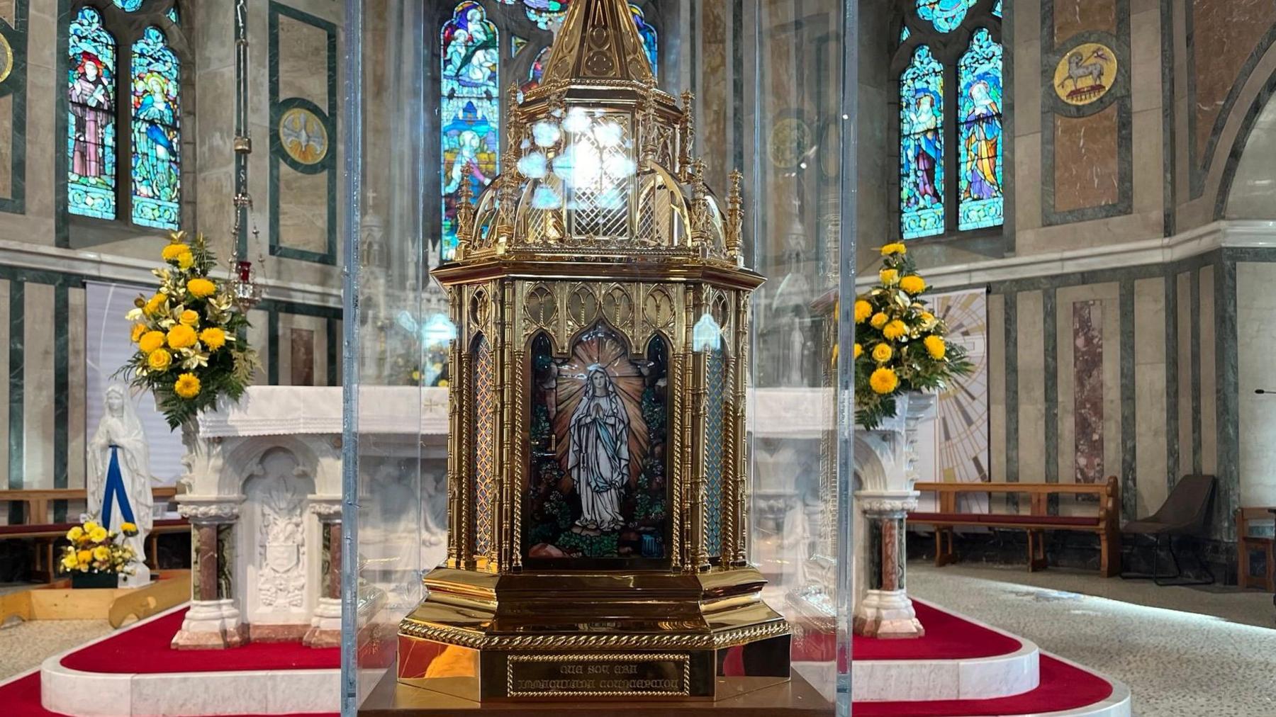 A gold decorative  reliquary  holding the relics of St Bernadette inside a perspex box in front of the altar of the Holy Cross Church